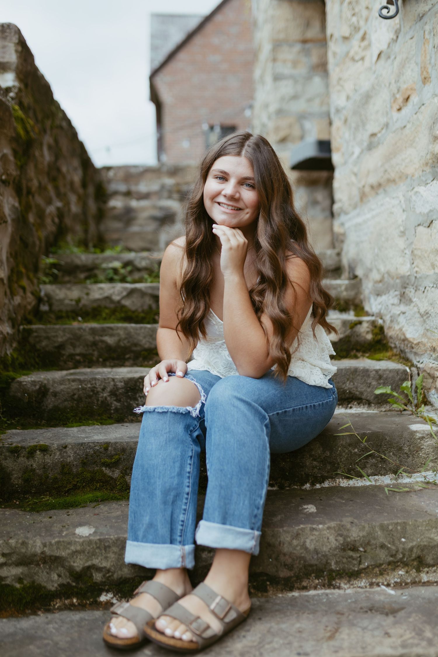 Young woman with long wavy hair sits smiling on stone steps, wearing white top, ripped jeans, and Birkenstock sandals.