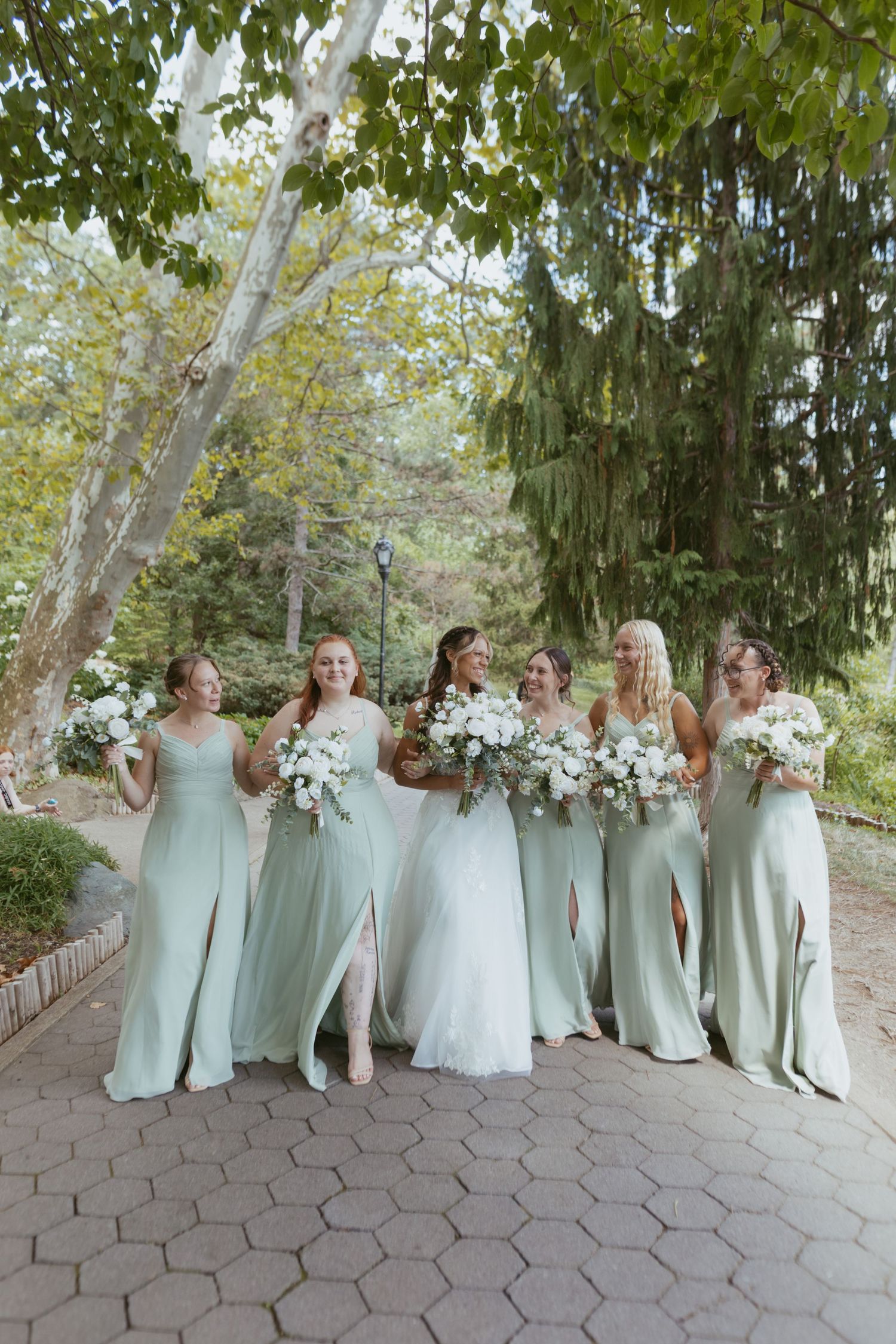 Bride in white lace gown walks with five bridesmaids in sage green dresses holding white bouquets in a garden.