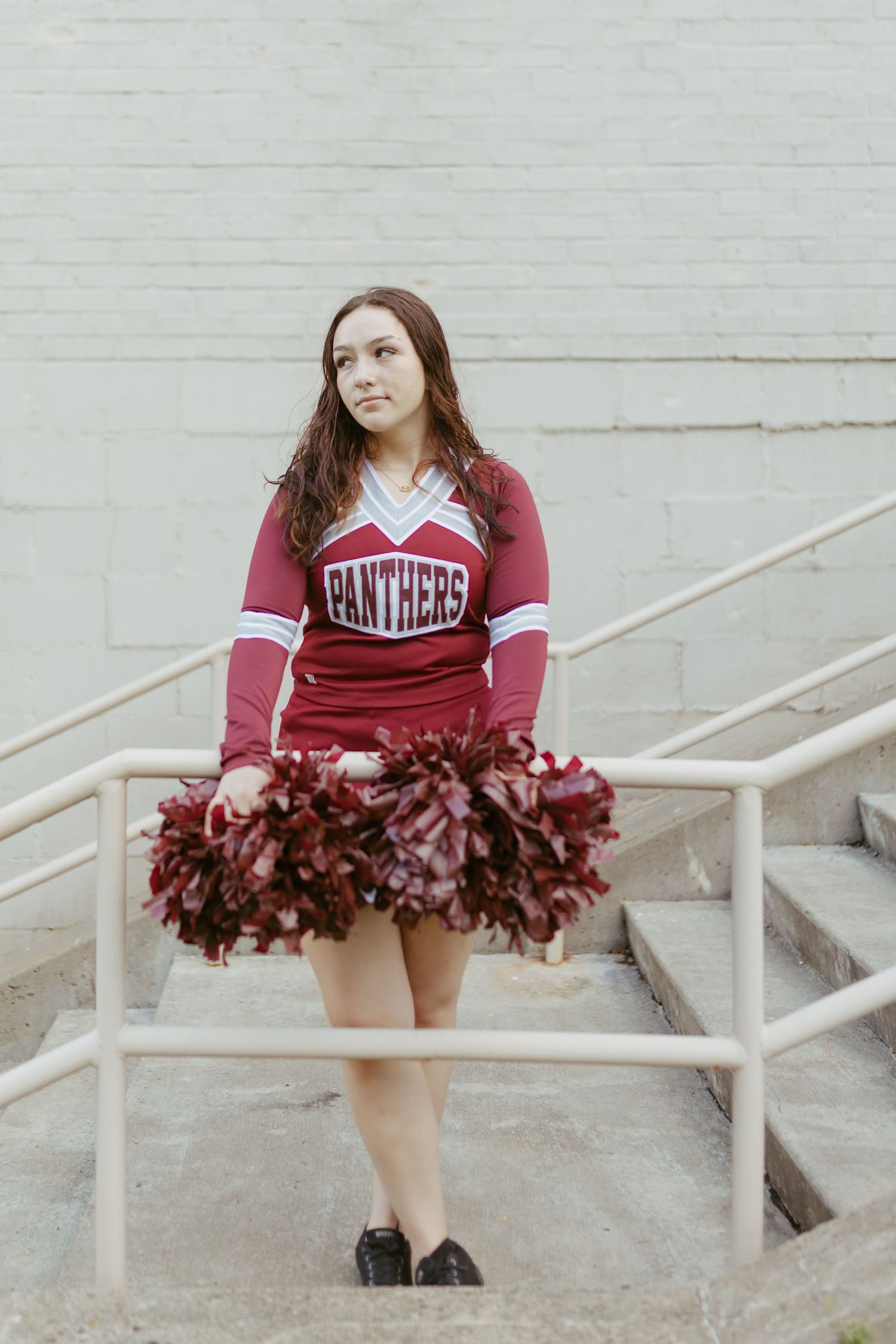 A cheerleader in a red Panthers uniform holds maroon pom-poms while standing on outdoor stairs.