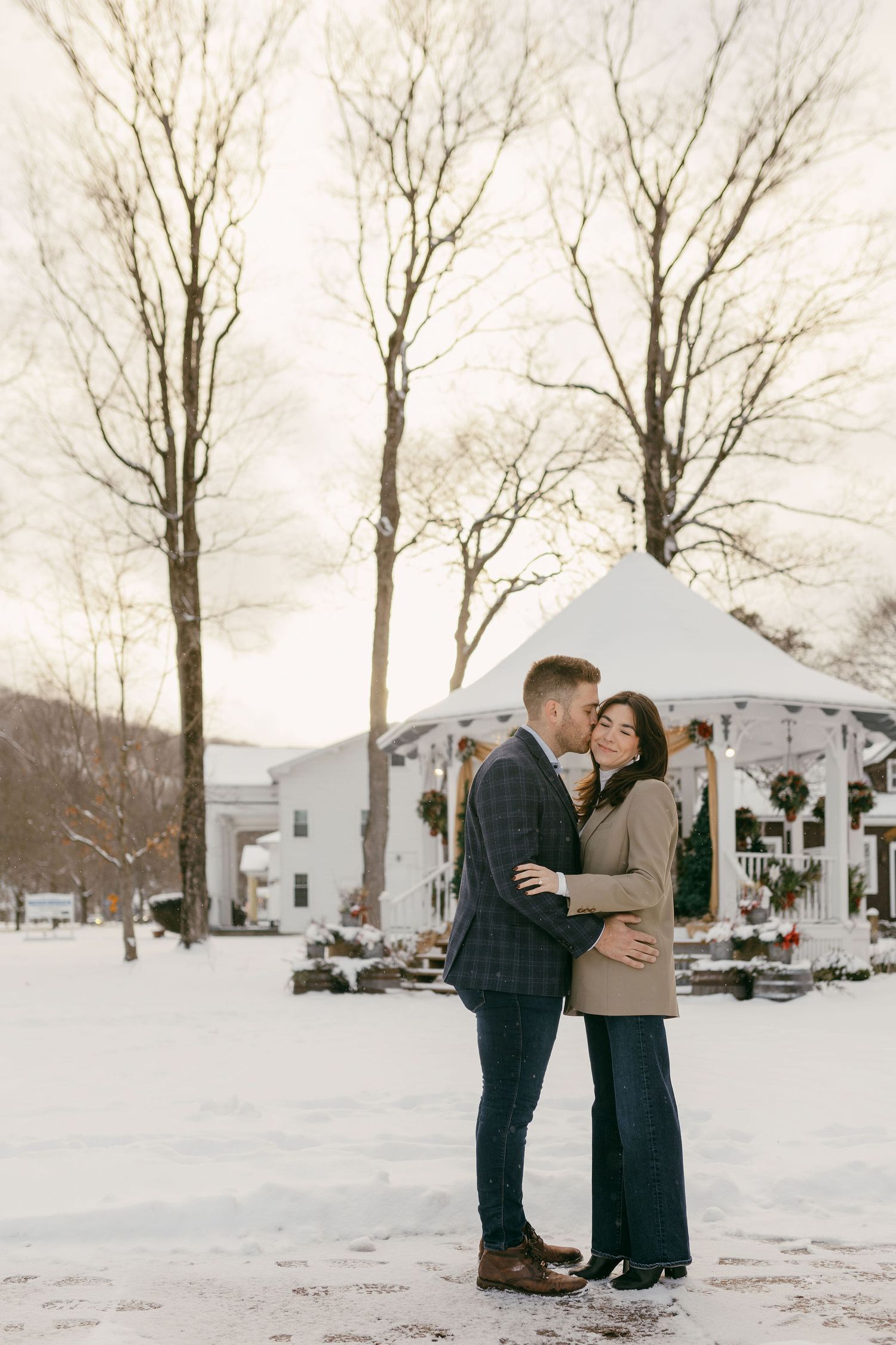 A couple embraces in front of a white gazebo on a snowy winter day.