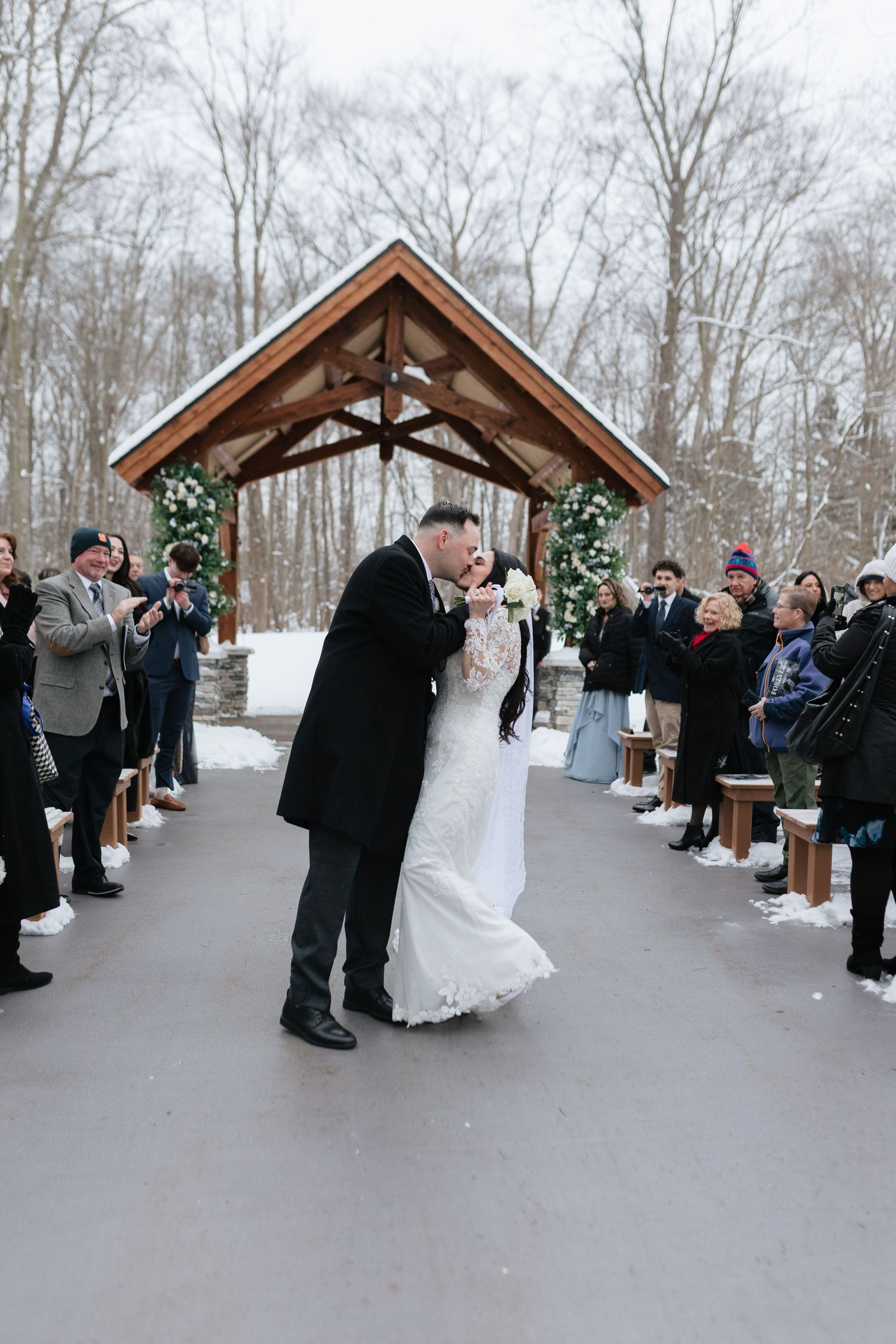 A winter wedding ceremony takes place outdoors under a wooden pavilion with snow-covered trees in the background.