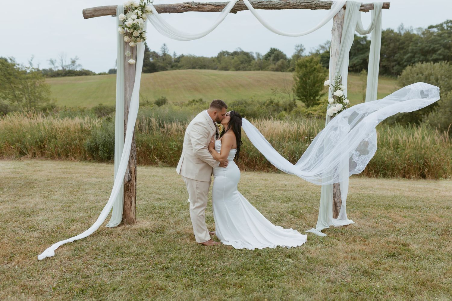 Couple sharing a romantic kiss under a rustic floral arch with flowing white drapes at an outdoor wedding ceremony.