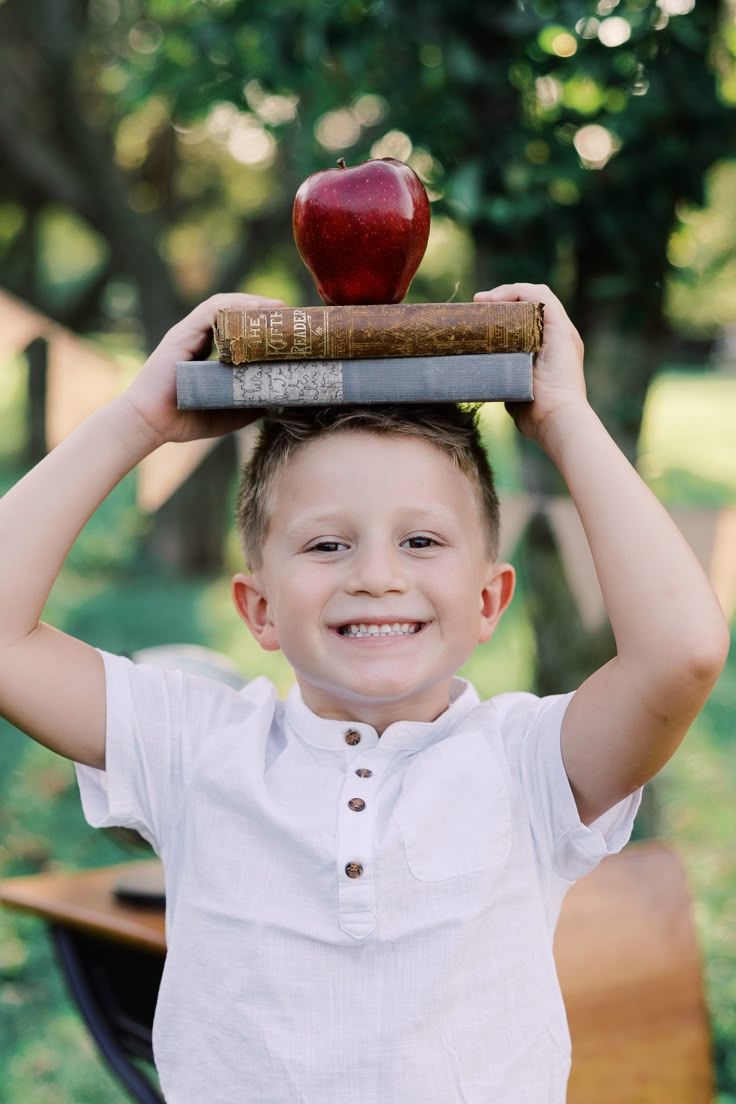 Smiling boy in white shirt holding stack of old books with red apple on top above his head outdoors.