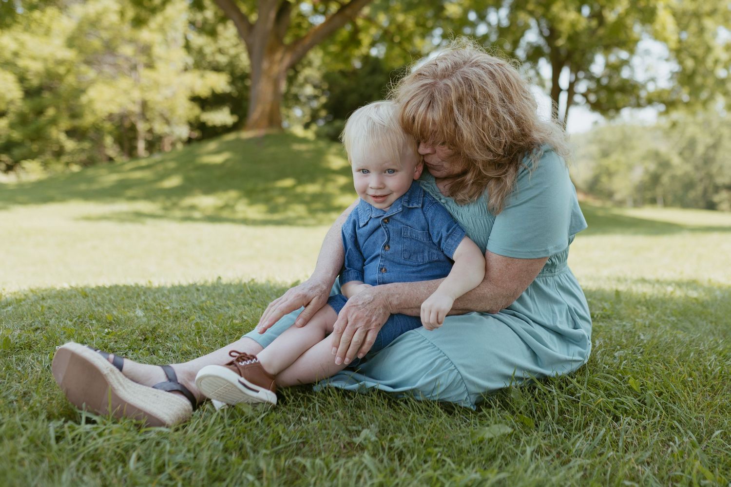 Grandmother with curly hair sits on green grass hugging a toddler in a blue outfit in a sunny park.
