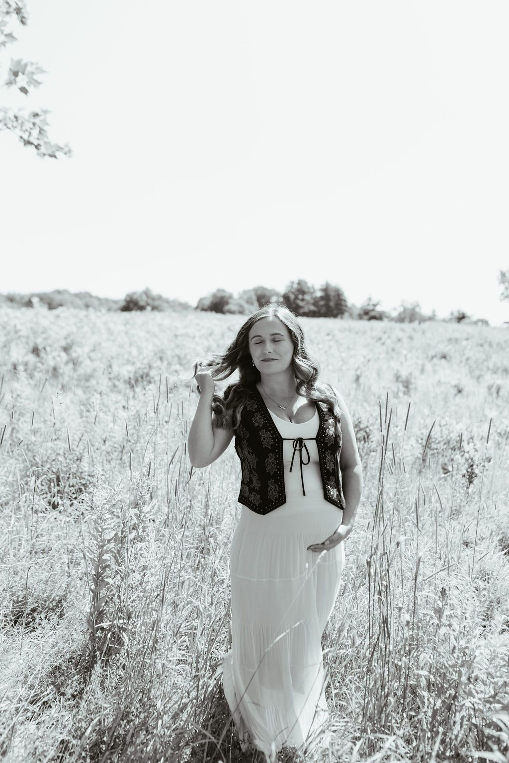 Black and white photo of a pregnant woman in a white dress and dark corset standing in a field holding a glass.