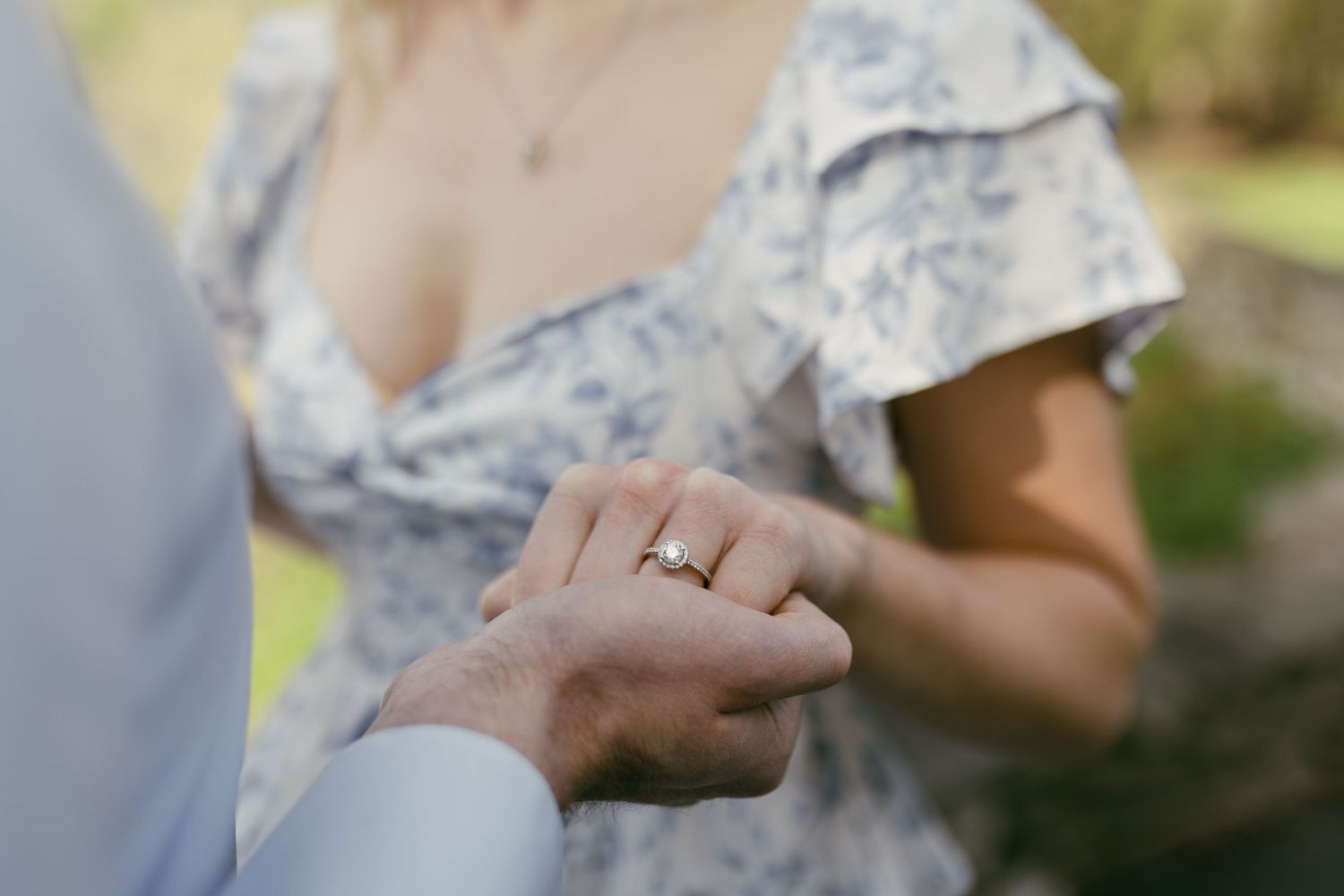 Close-up of a woman in a floral dress showing off her diamond engagement ring while holding hands with her partner outdoors.