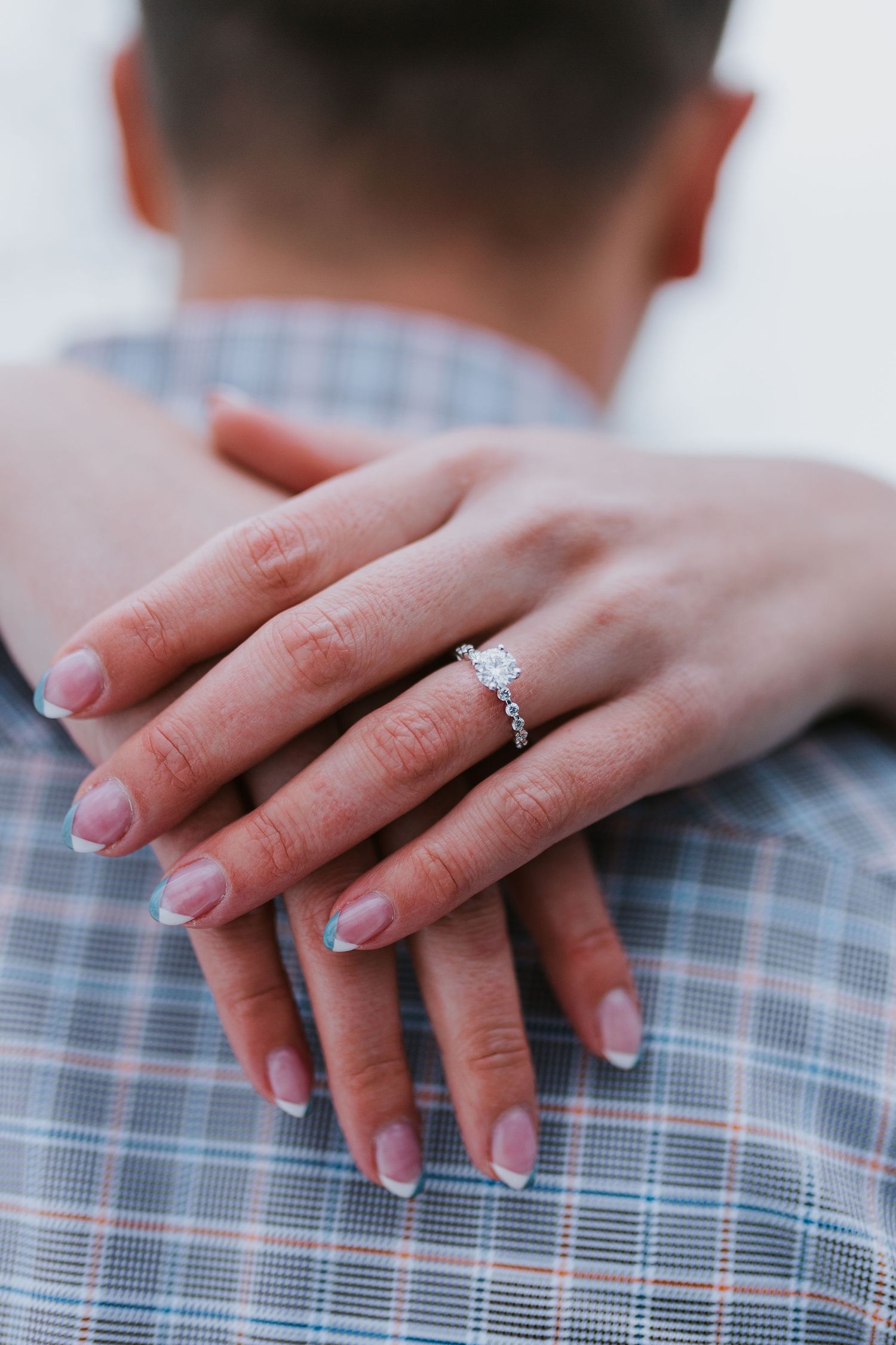 Close-up of a woman's hand displaying a diamond engagement ring while embracing a man in a plaid shirt.