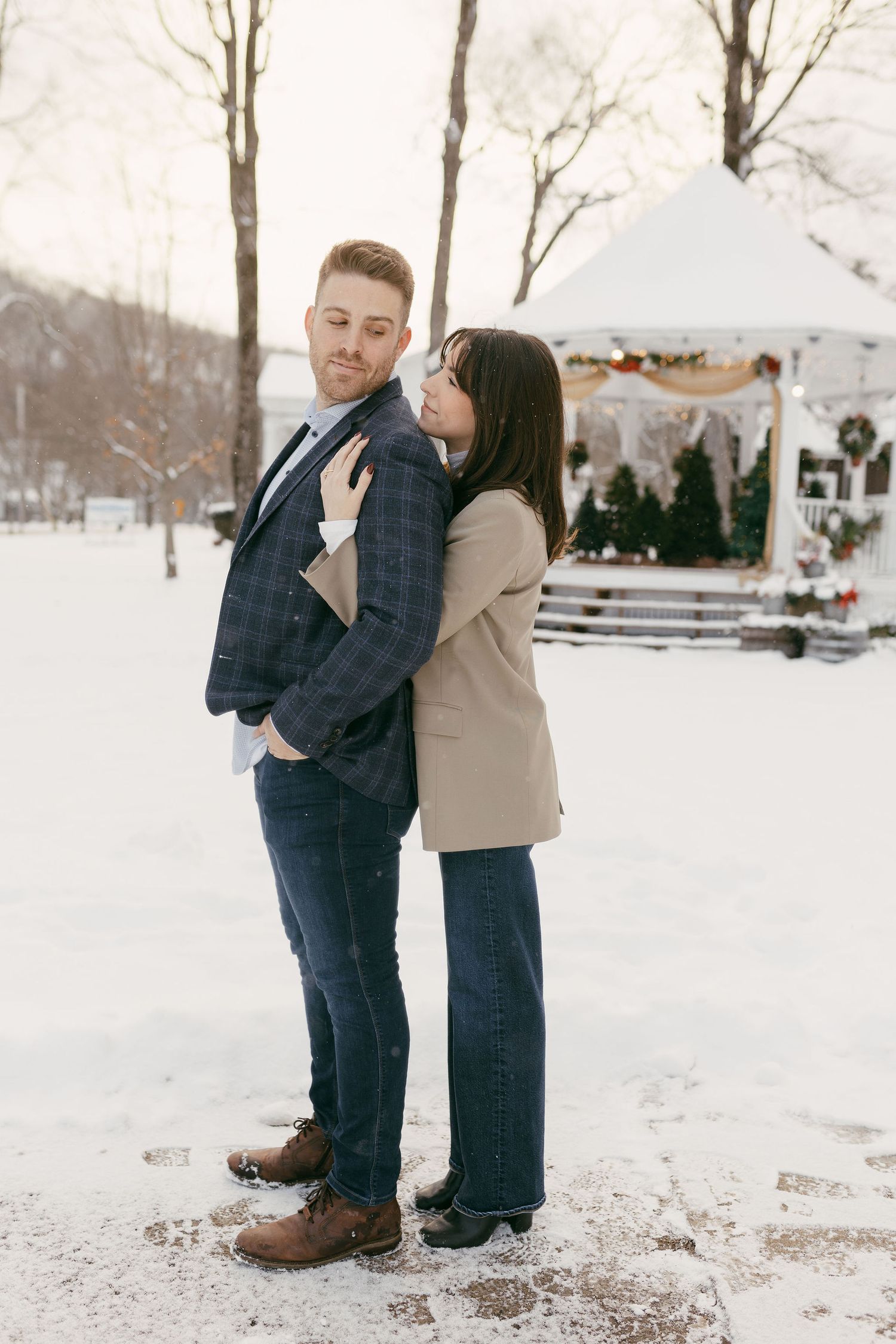 Couple embracing in snowy park, man in plaid blazer and jeans, woman in beige jacket, festive gazebo background.
