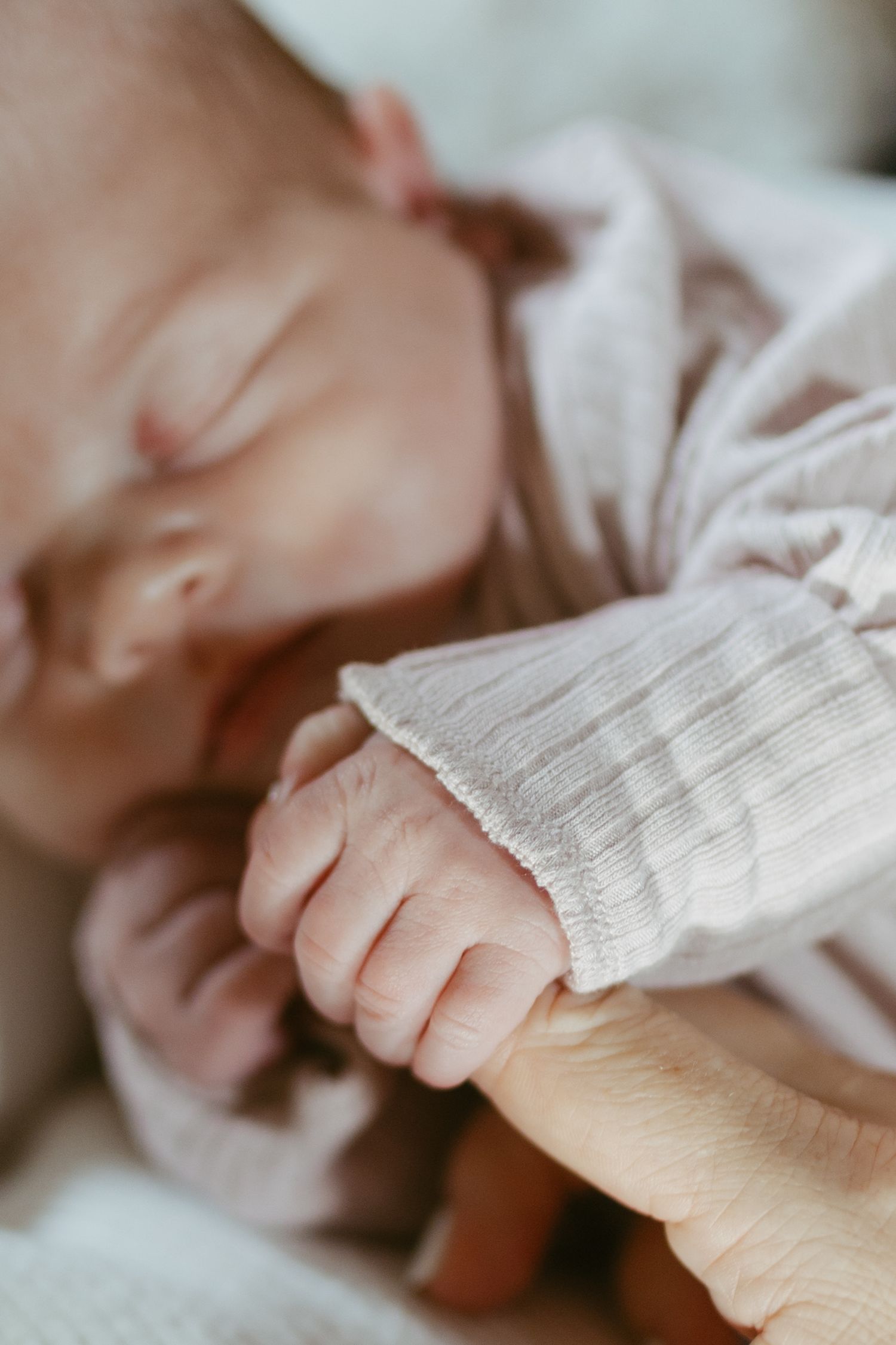 Sleeping newborn baby in ribbed onesie gently gripping an adult finger with tiny hand.