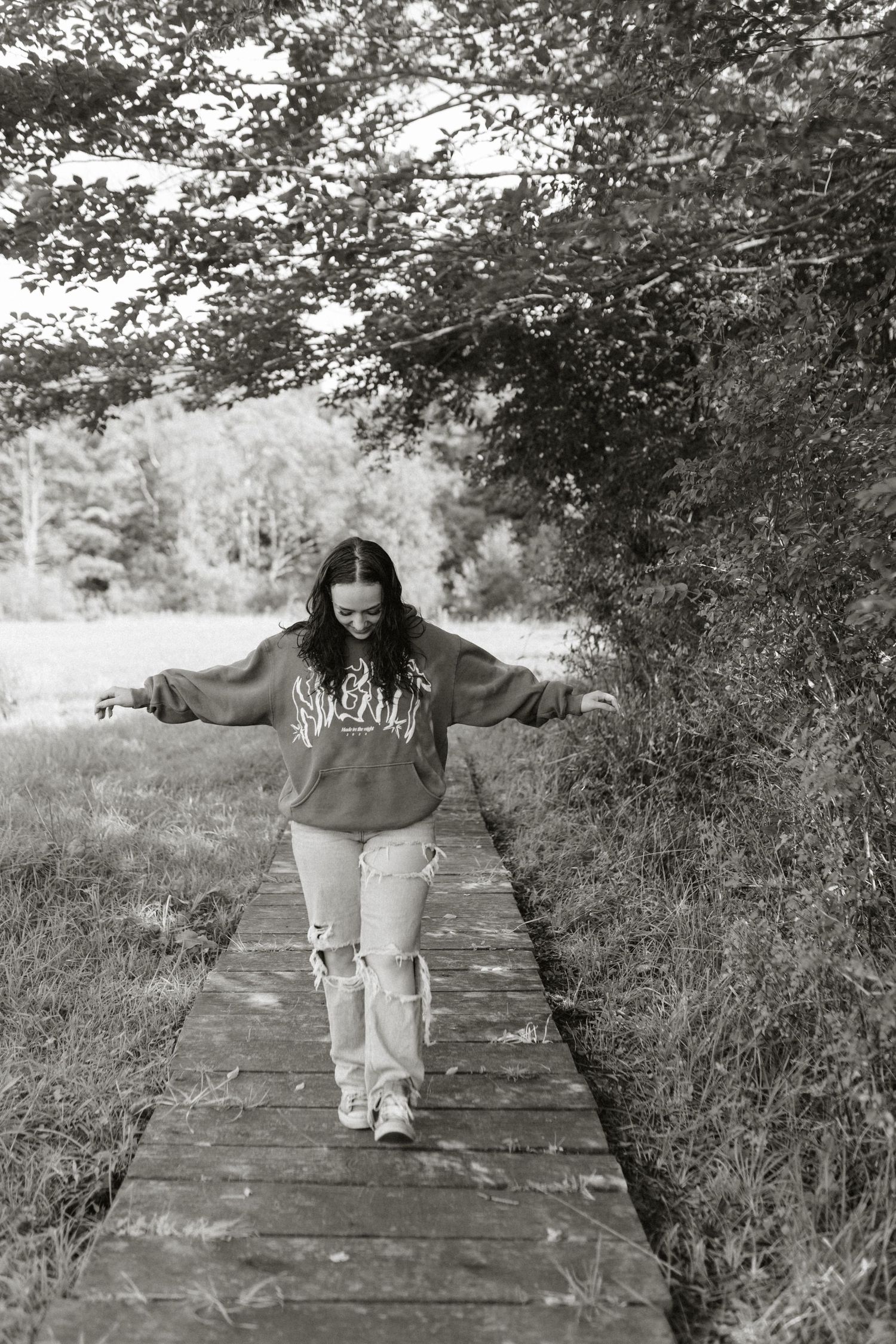 Young woman balancing on wooden boardwalk path in ripped jeans and graphic sweatshirt, black and white outdoor photo.