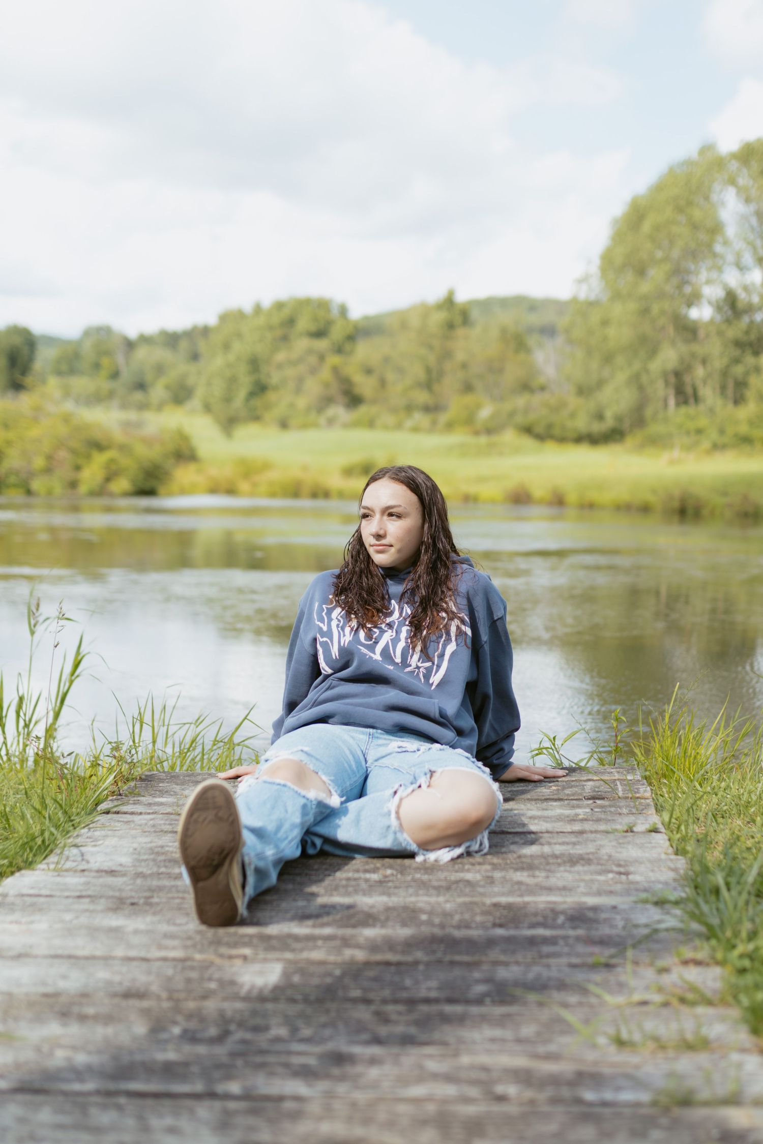 Young woman in ripped jeans and gray sweatshirt sitting on wooden dock by a serene lake surrounded by lush greenery.