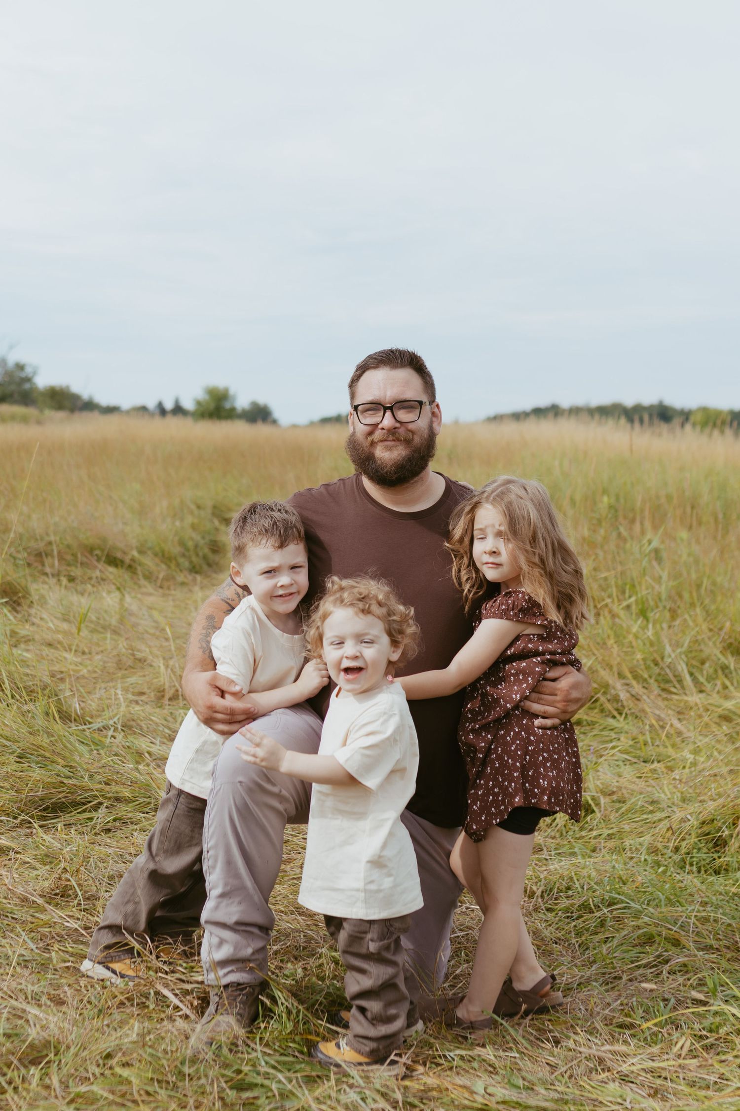 A bearded father with glasses poses with three young children in a golden field, smiling for a family portrait.