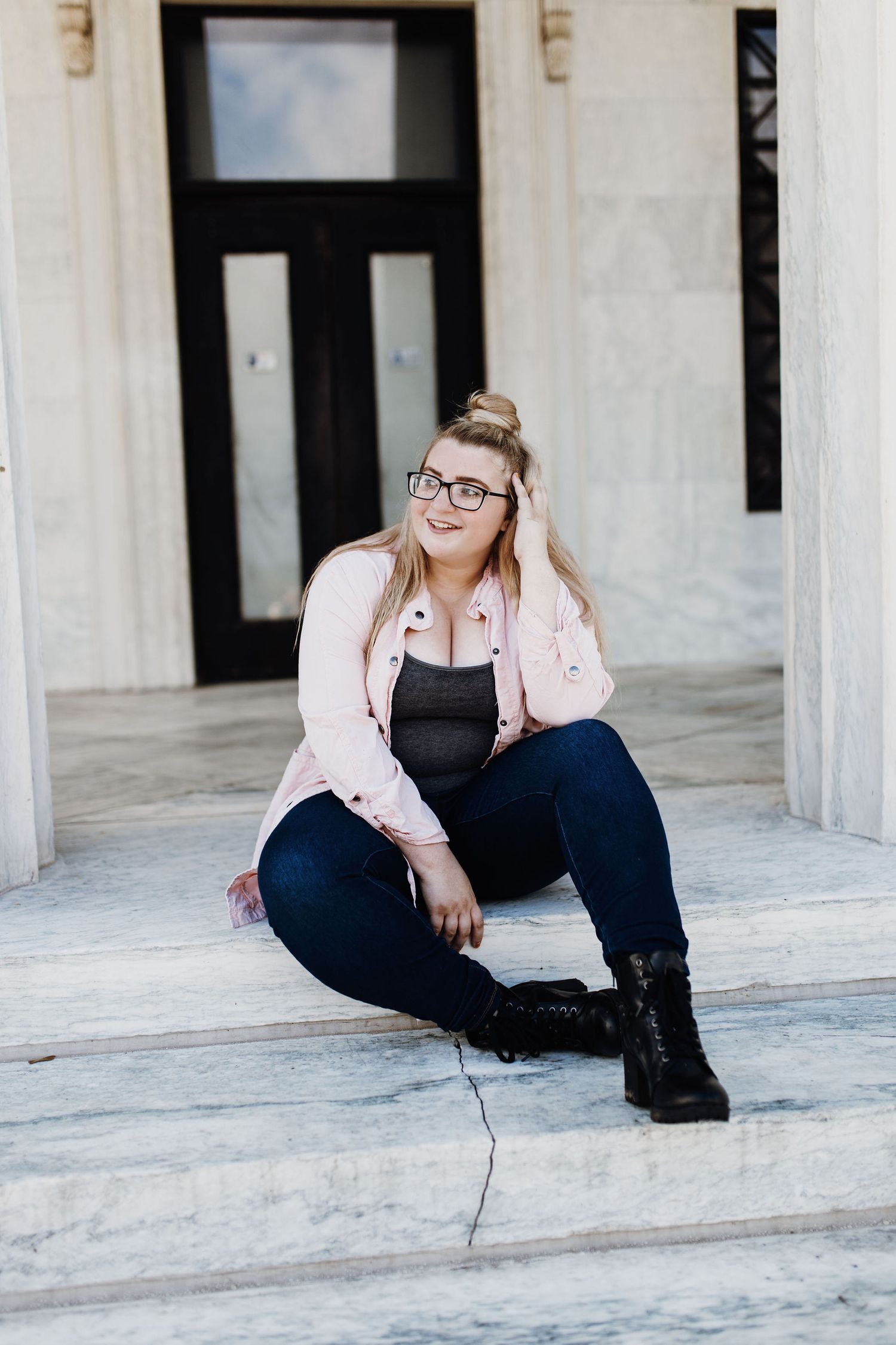 Young woman in pink jacket, jeans, and boots sitting on marble steps in front of a building.