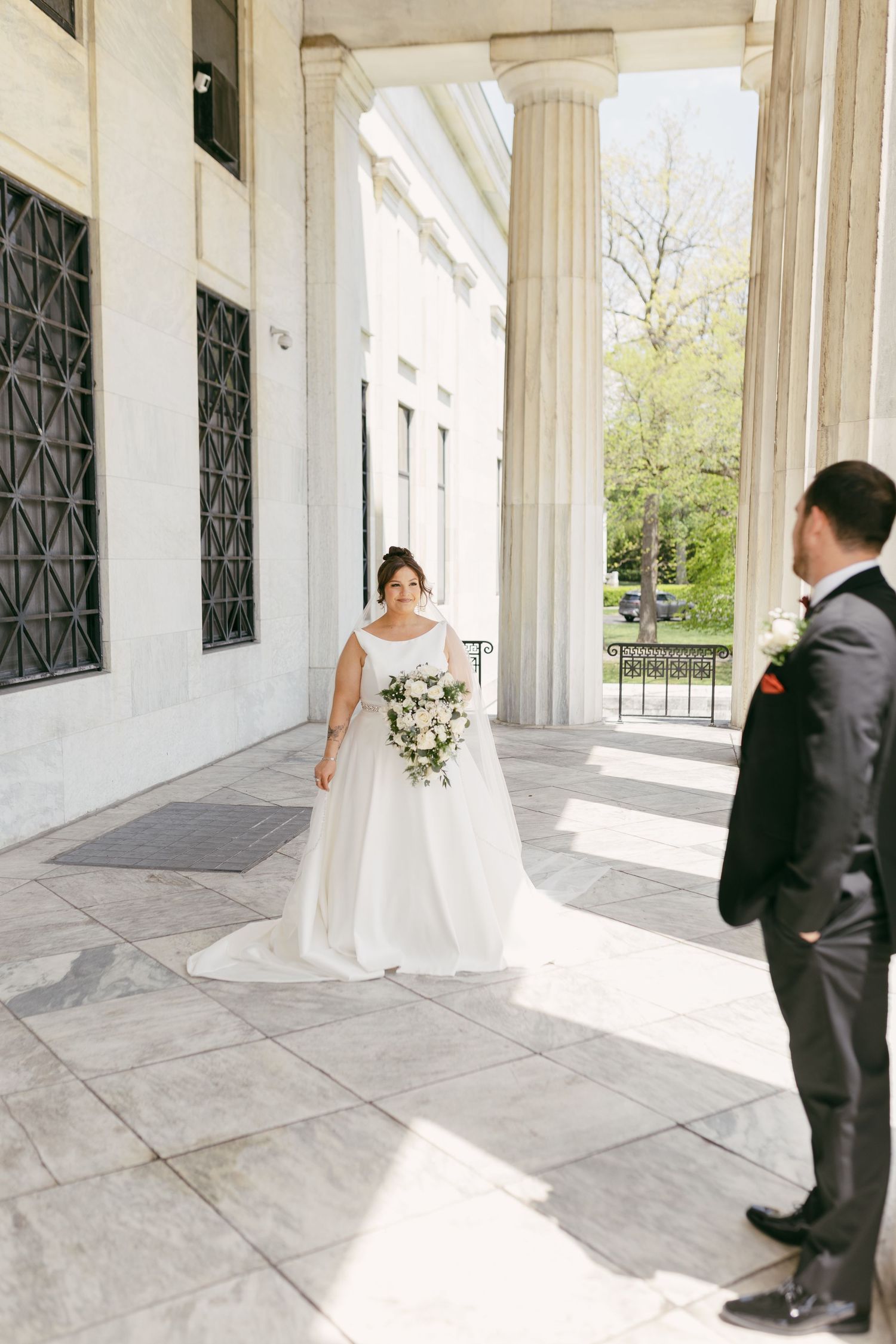 Bride in white gown holds bouquet during first look with groom at elegant columned marble building.