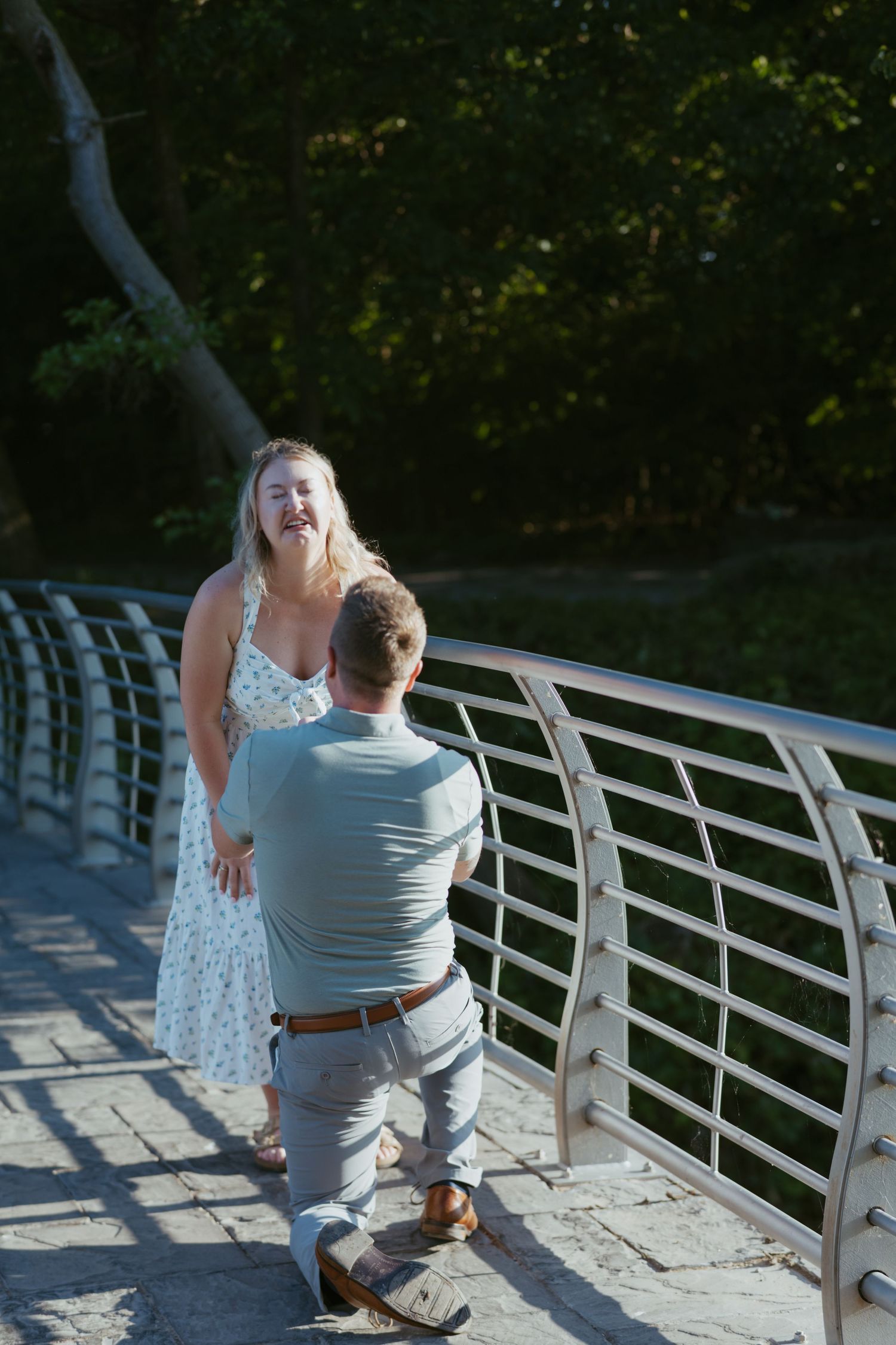 Man proposing on knees to surprised woman on a bridge in a sunny park setting.