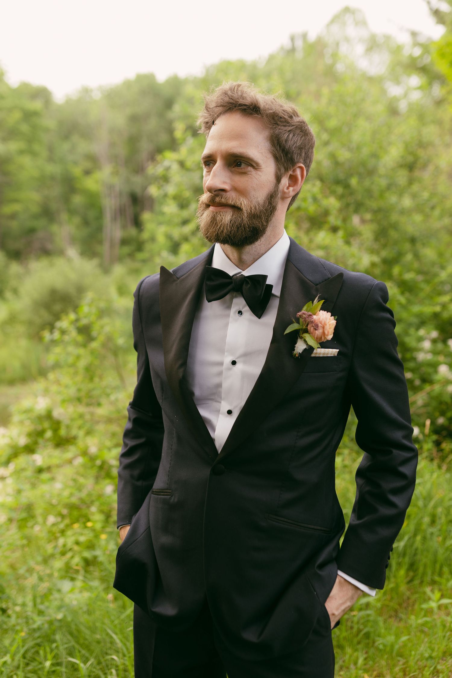 Groom in classic black tuxedo poses against natural greenery backdrop.