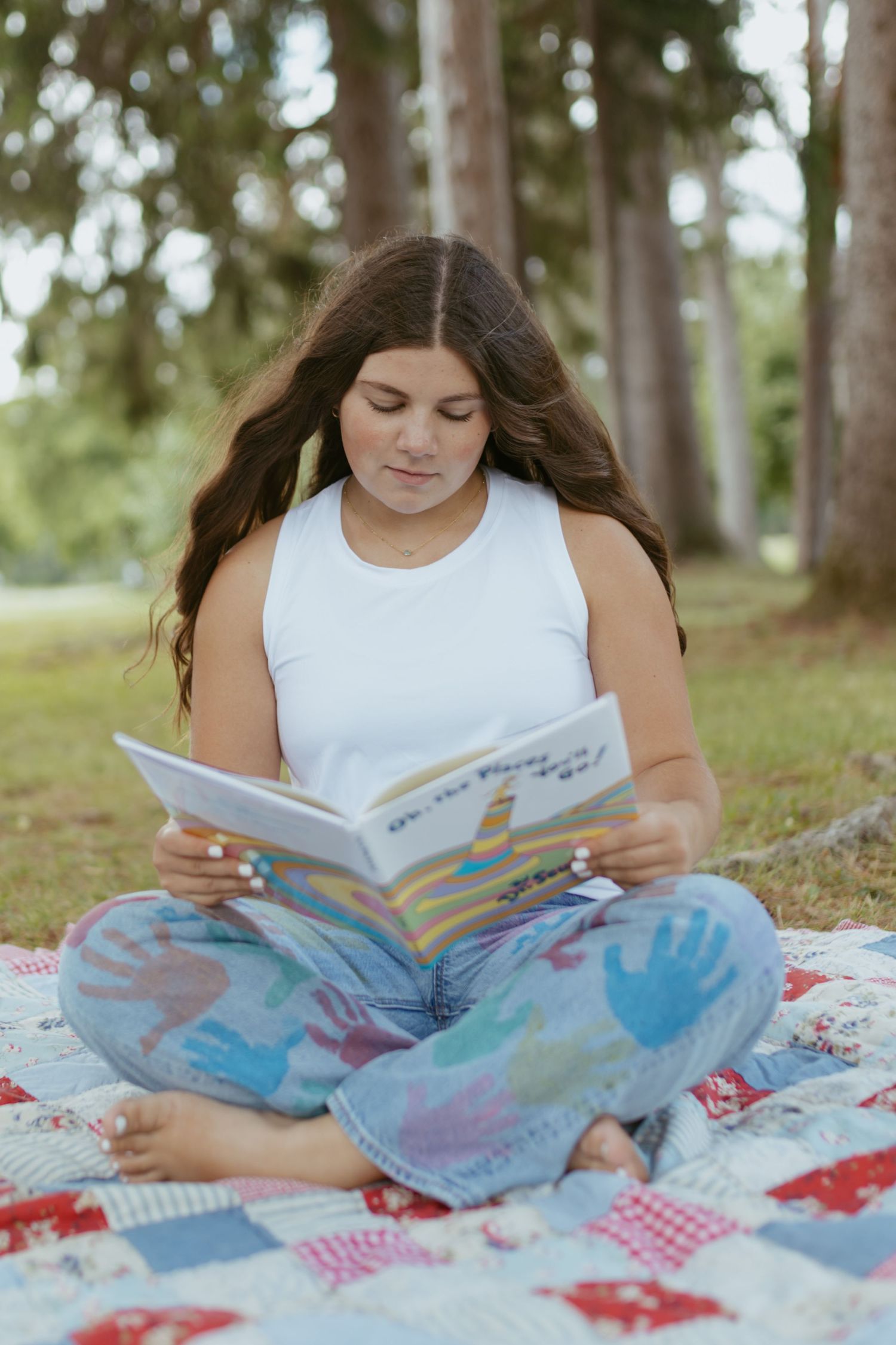 Young girl sitting cross-legged on a quilt outdoors, reading a colorful book among tall trees.