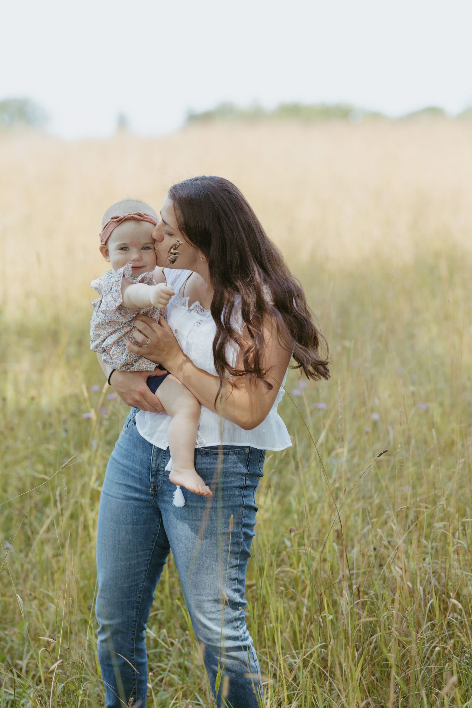 A mother kisses her baby girl while standing in a golden field, both wearing casual outfits on a sunny day.
