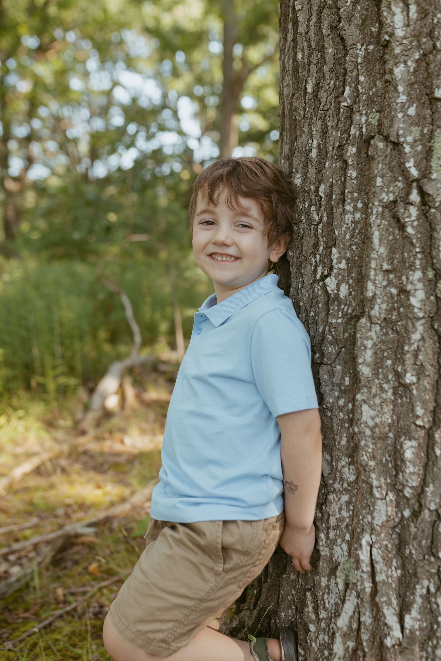 A smiling young boy in a light blue polo shirt and khaki shorts leaning against a large tree trunk outdoors.