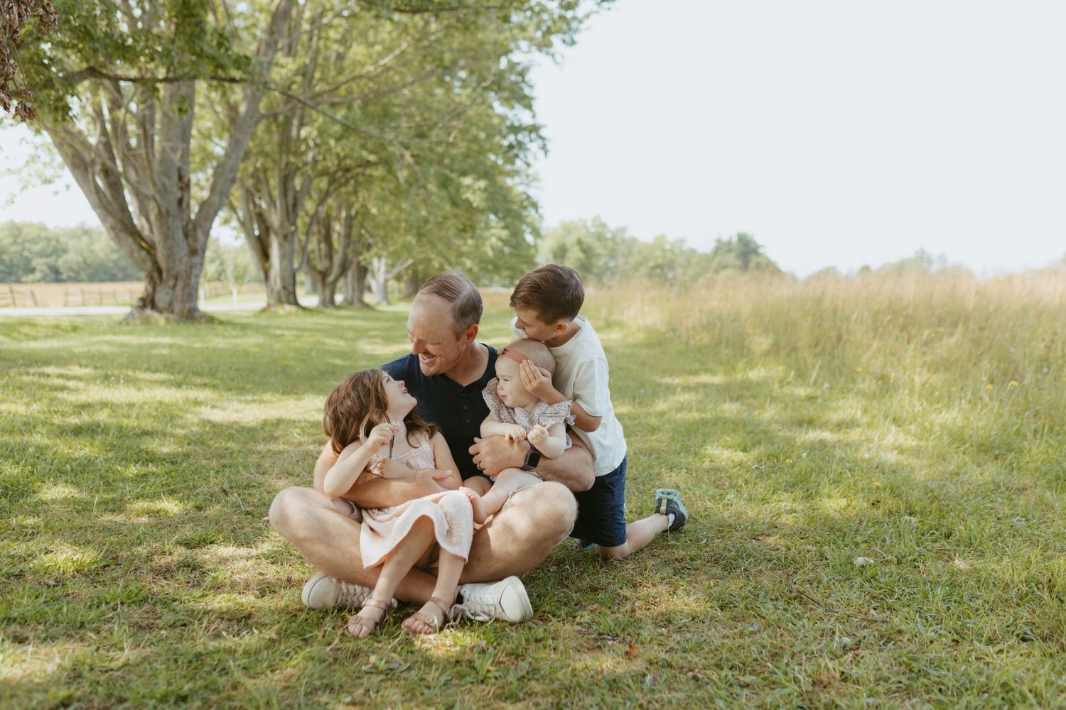 A father sits on grass with three young children in a sunny park, sharing a warm family moment surrounded by trees.