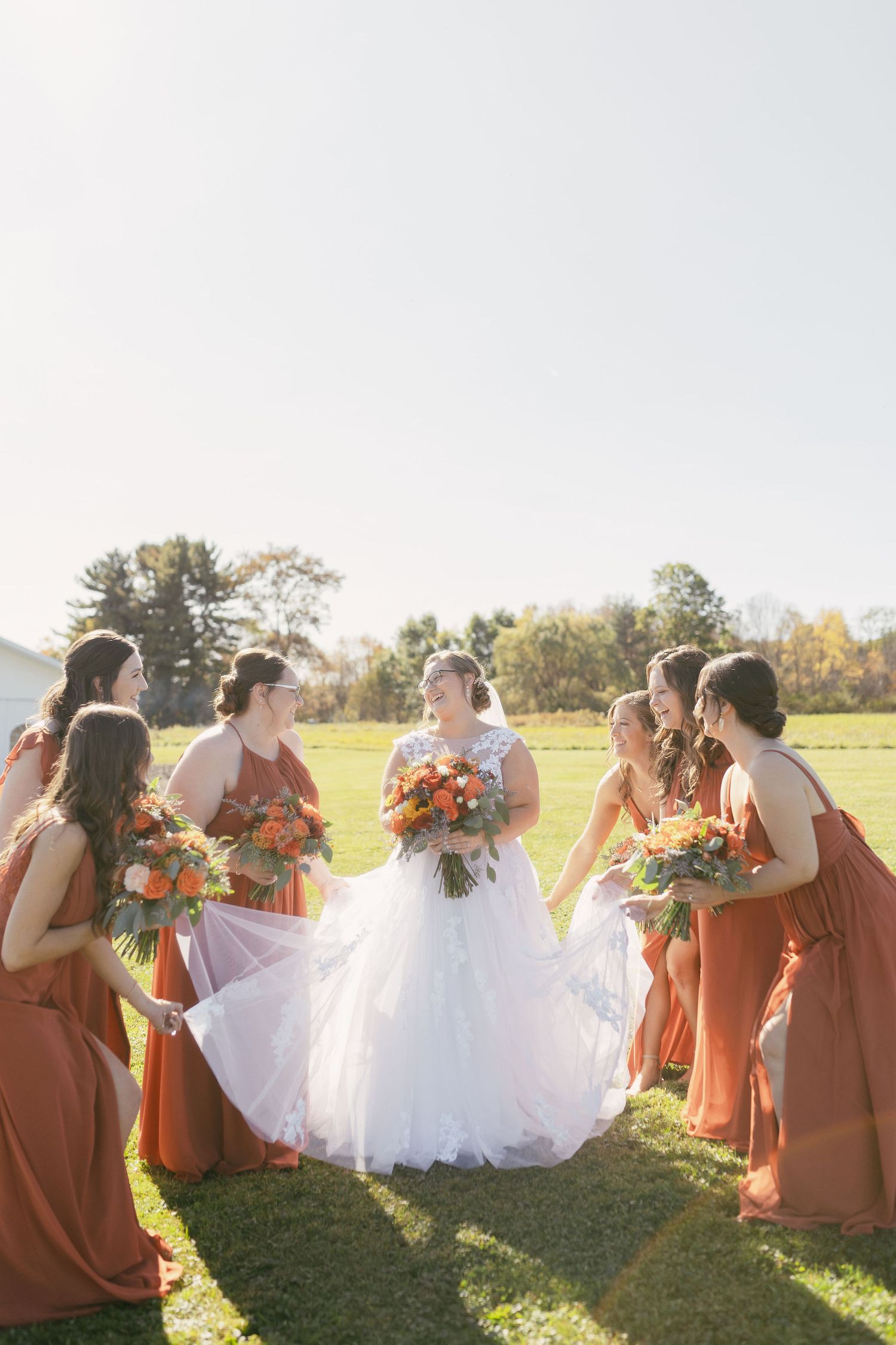 Wedding party in orange dresses poses on a sunny grassy hill with autumn trees in the background.
