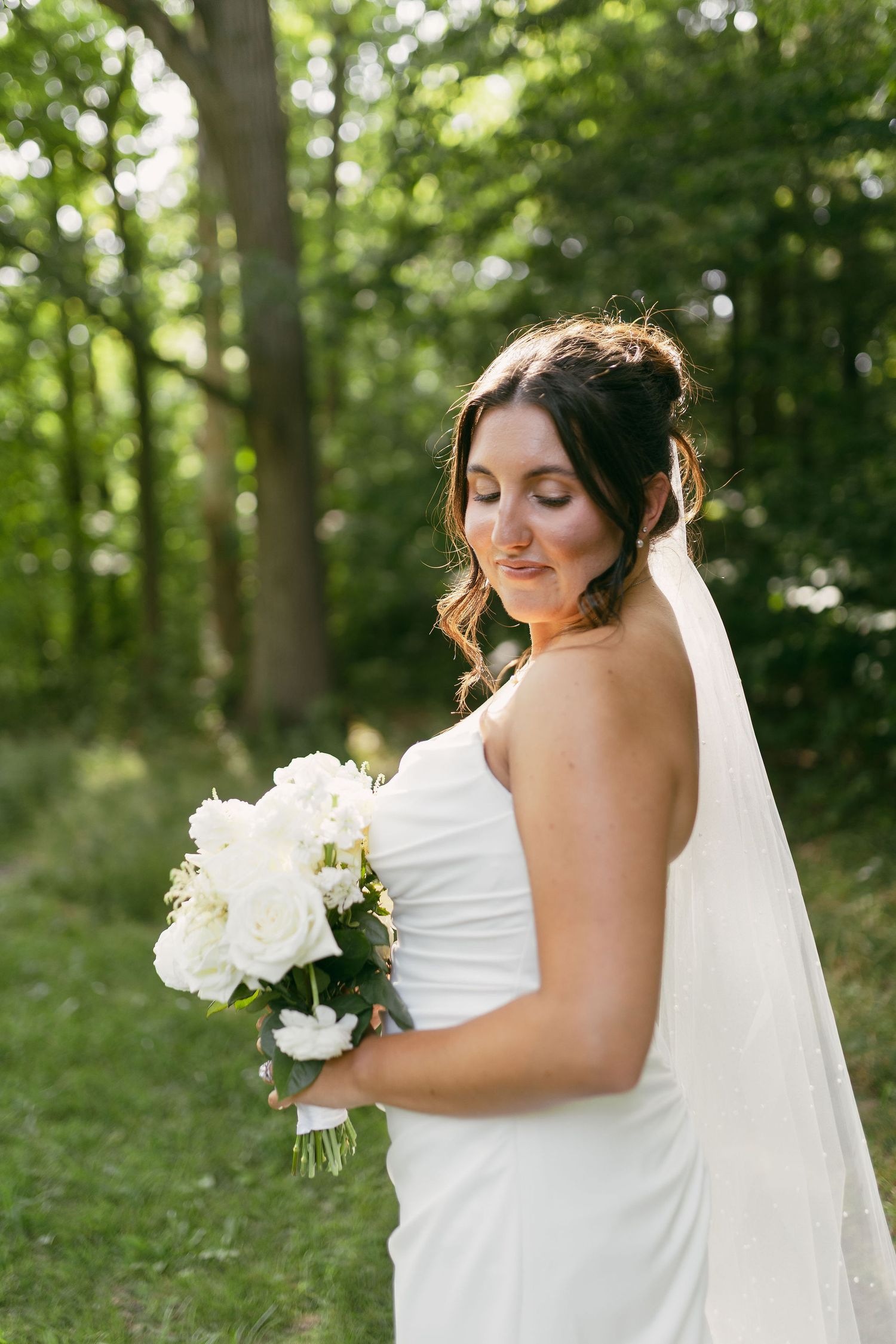 Bride in white dress holds classic white bouquet while posing in a wooded setting.
