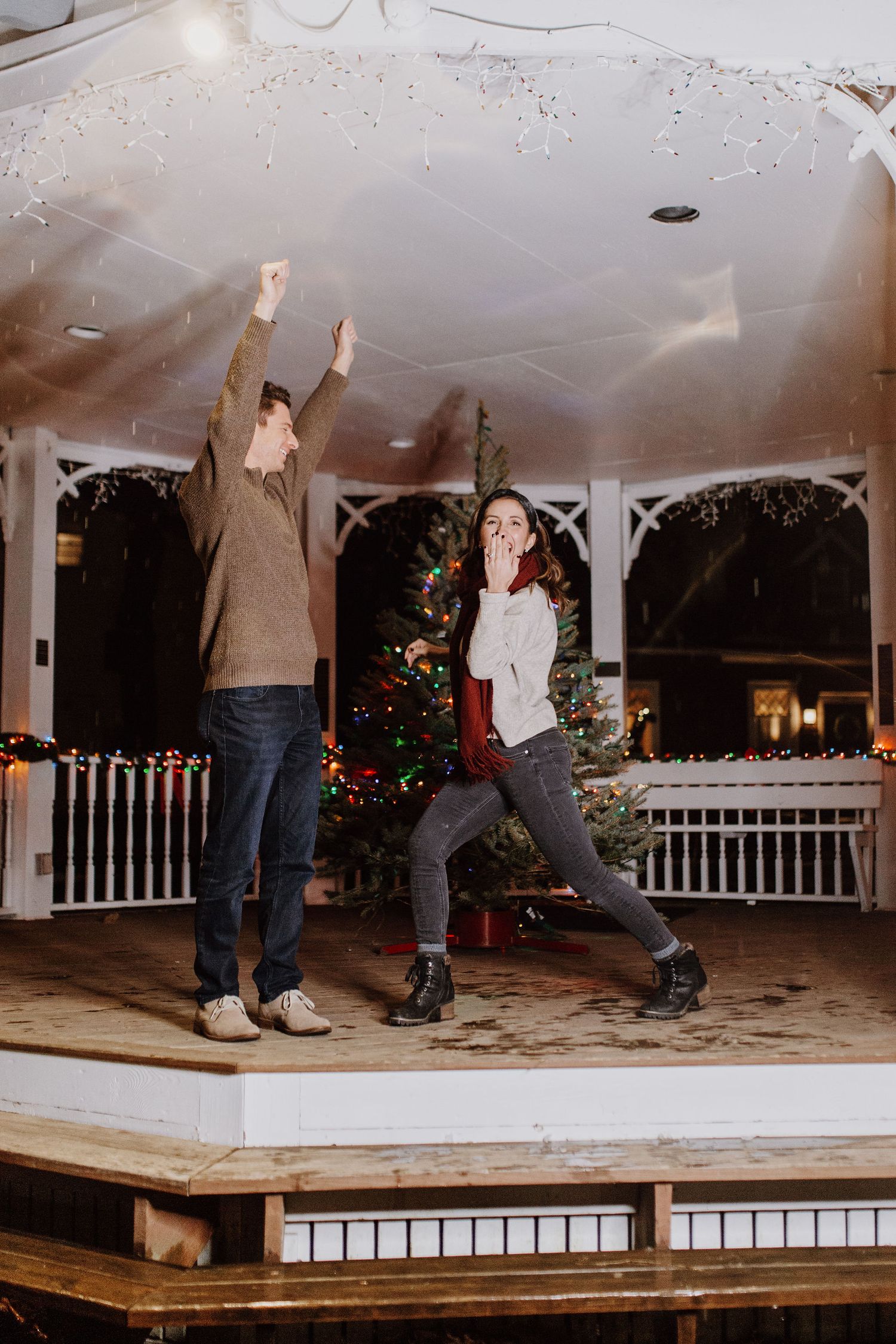 Couple celebrating joyfully in a decorated gazebo with Christmas tree lights at night.