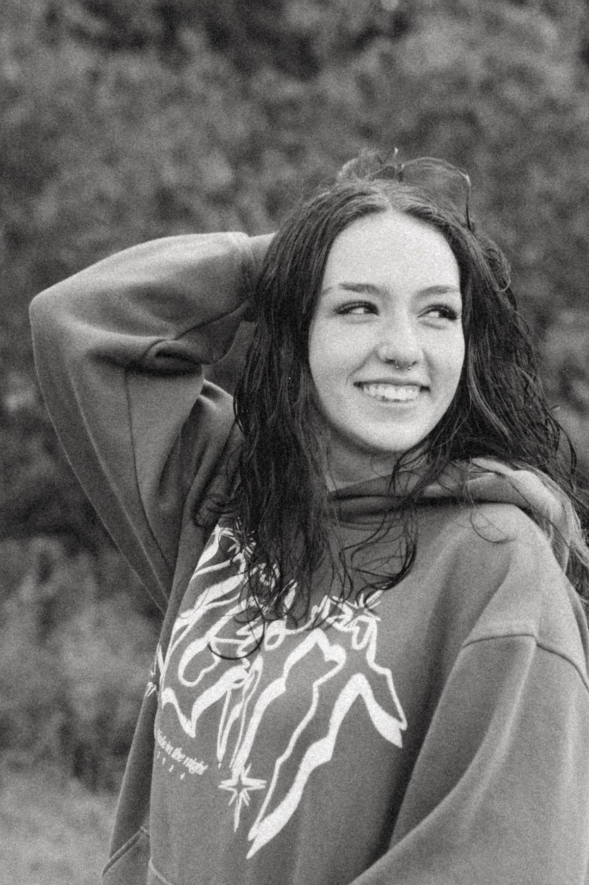 Black and white photo of a smiling young woman with long hair wearing a graphic hoodie outdoors.