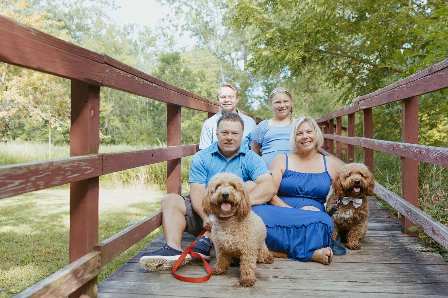 Family of four in blue outfits poses with two golden doodle dogs on a wooden bridge surrounded by trees.