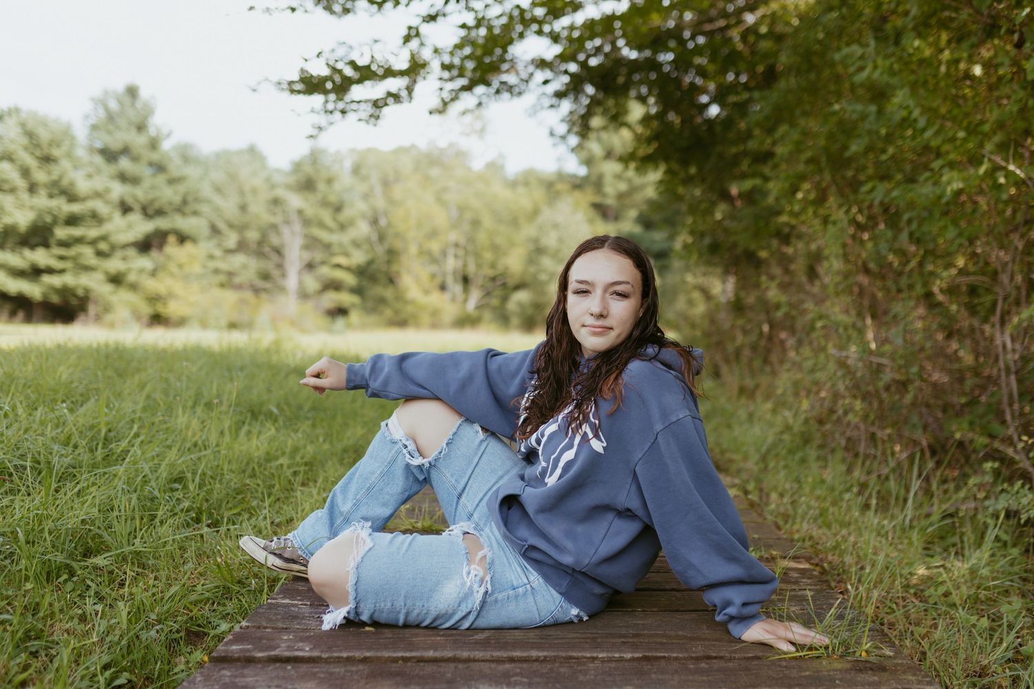 Young woman in ripped jeans and blue hoodie sitting on wooden dock surrounded by lush green meadow and trees.