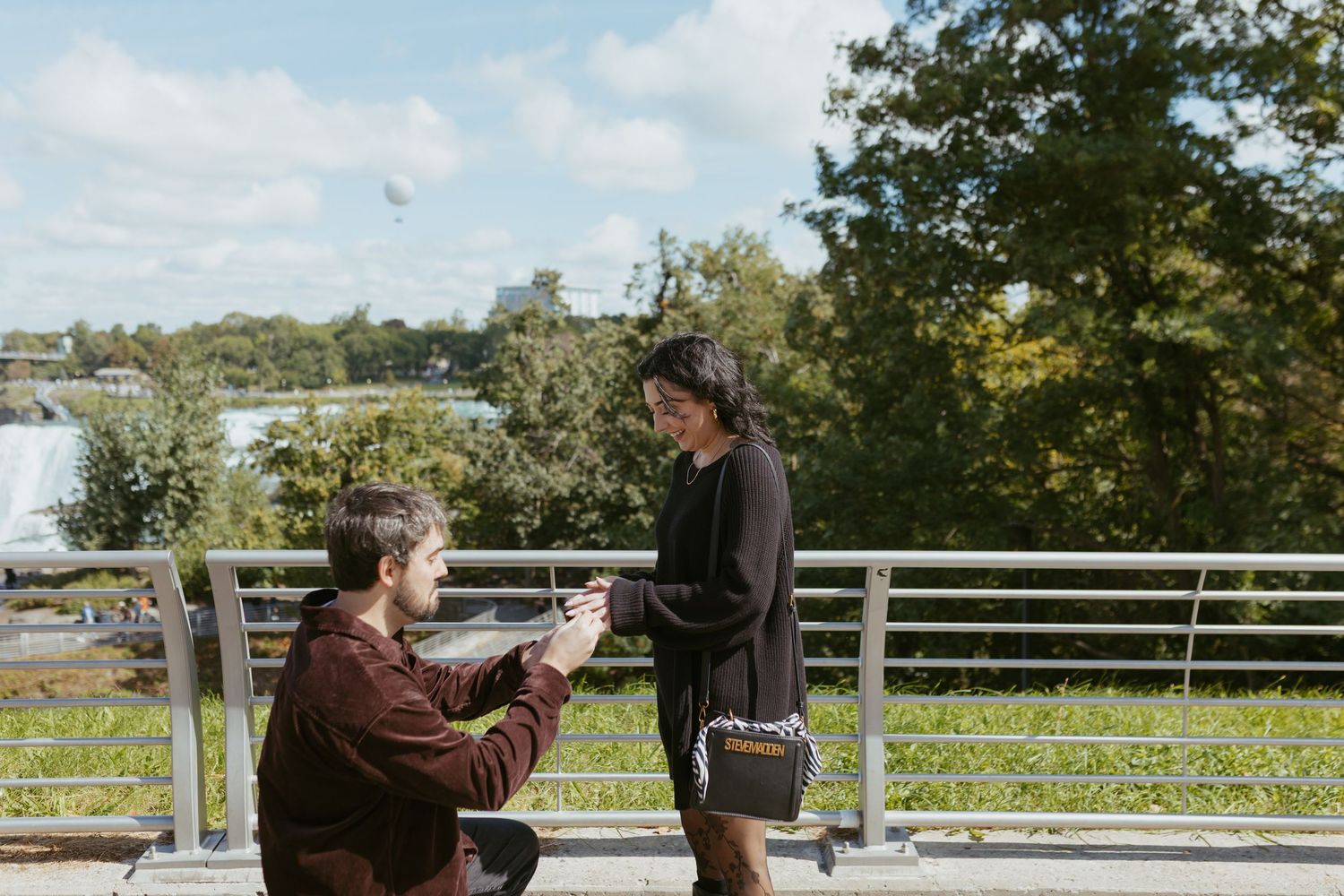 Man kneeling on one knee proposing to partner on an outdoor terrace with lush green trees in the background.