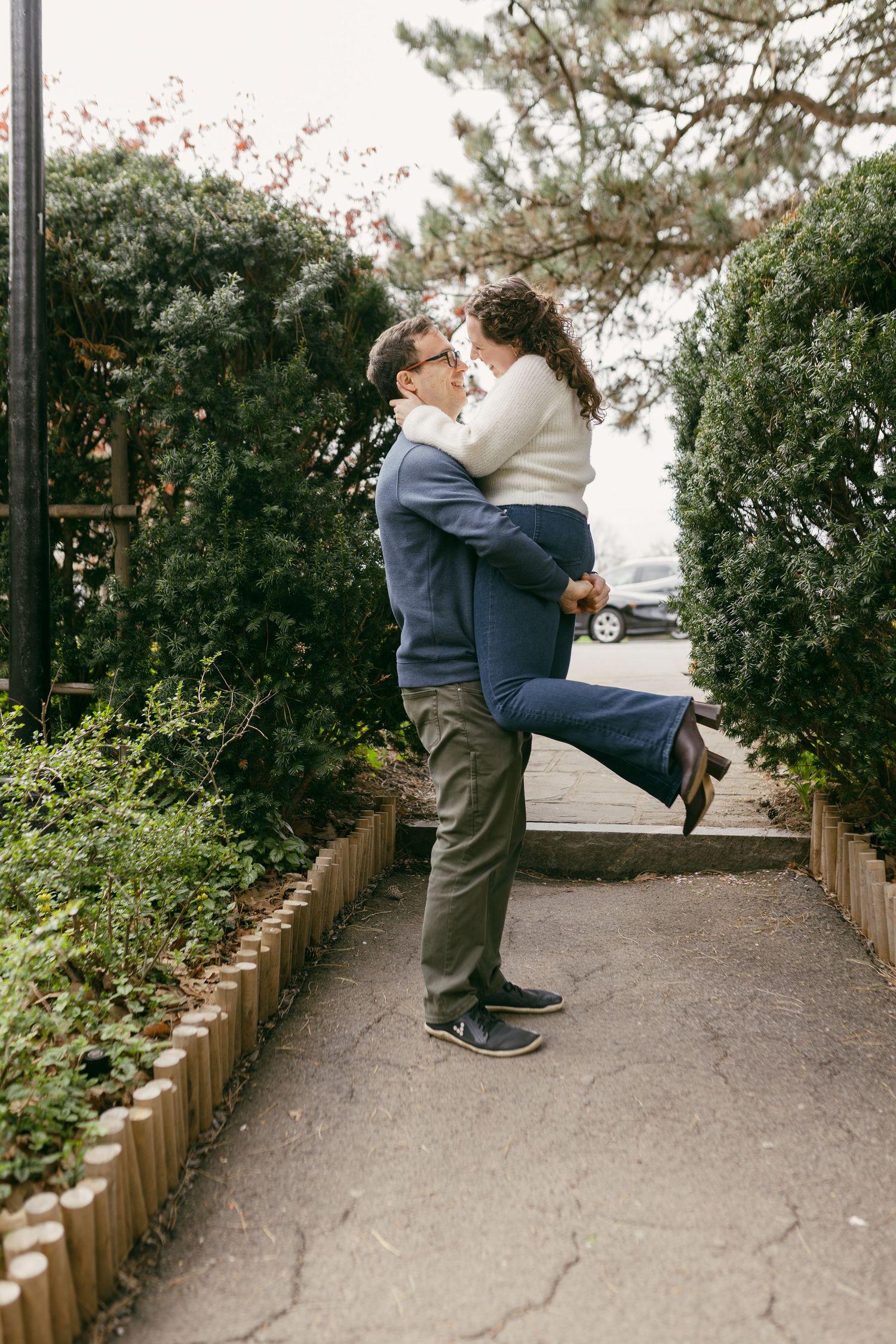 A couple shares a romantic embrace on a garden pathway, the man lifting the woman as they smile at each other.