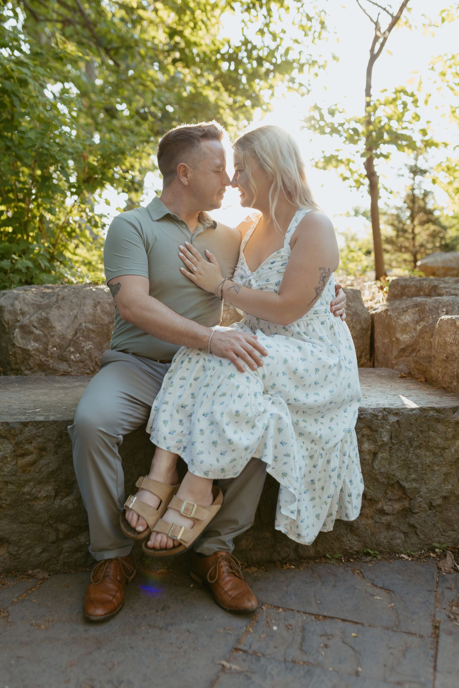 A couple shares a romantic moment sitting on stone steps surrounded by lush green trees in warm sunlight.