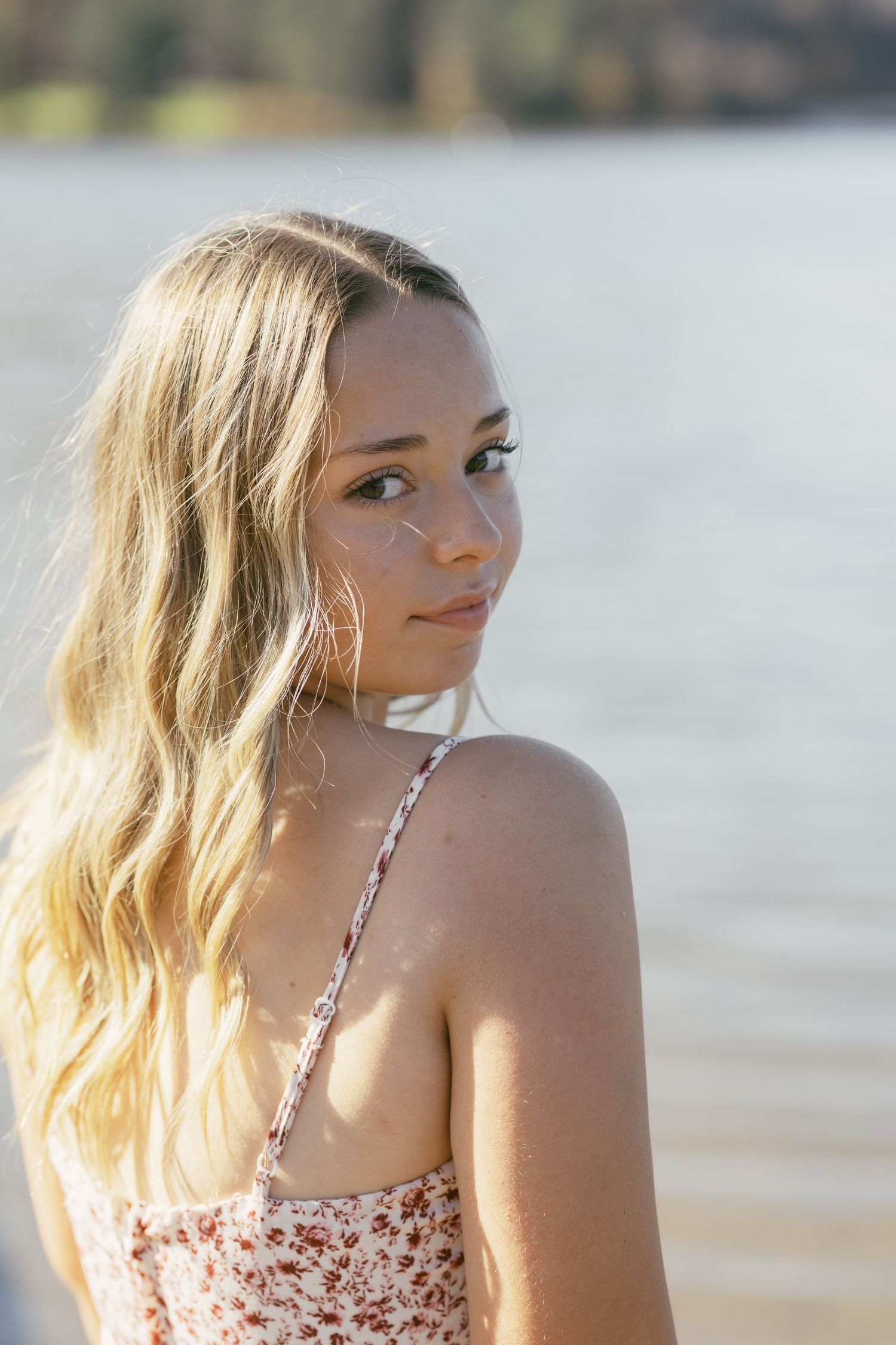 Blonde teen girl in floral sundress looks back over shoulder near sunlit lake.