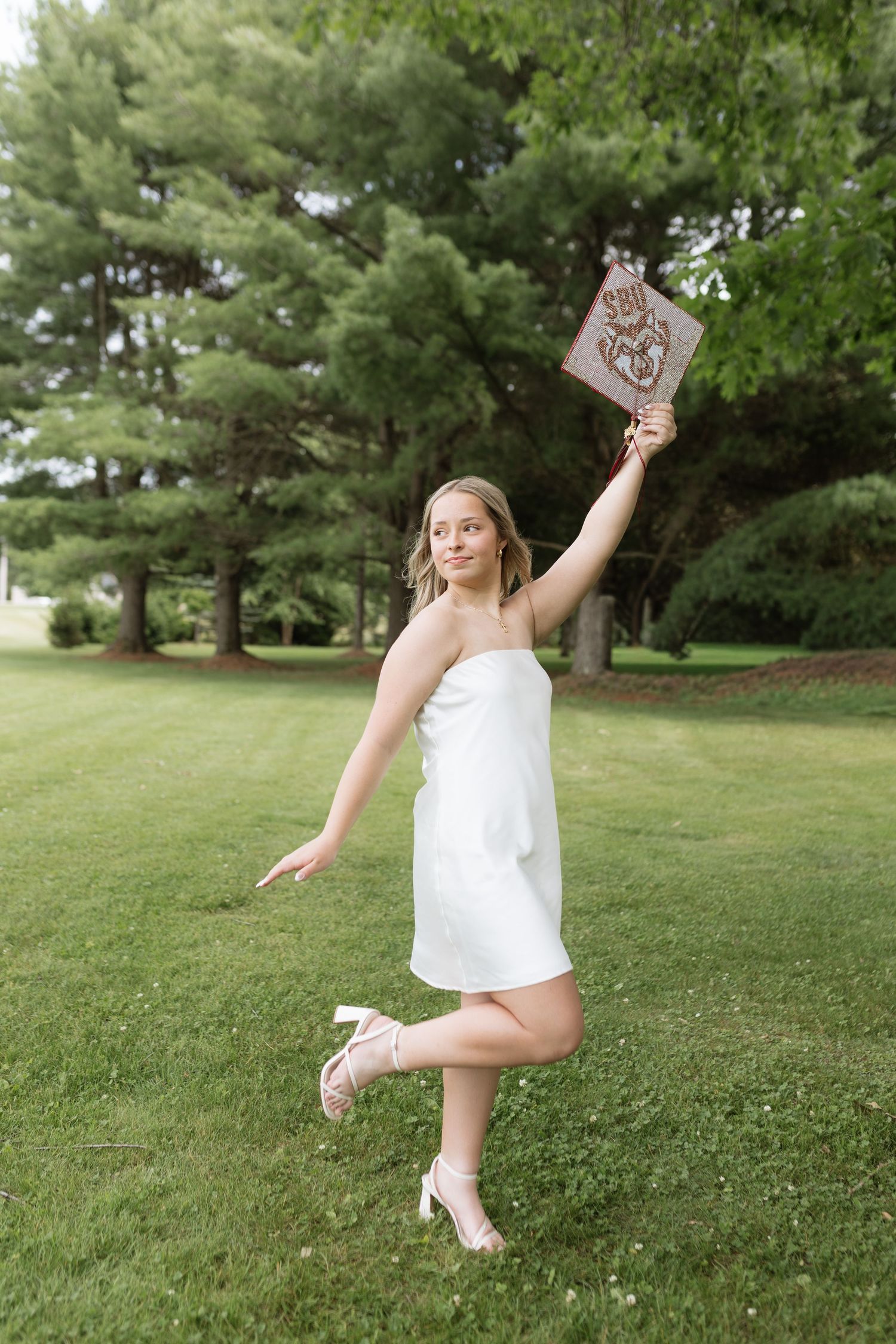Young woman in white dress holding diploma aloft on one leg in lush green park, celebrating graduation outdoors.