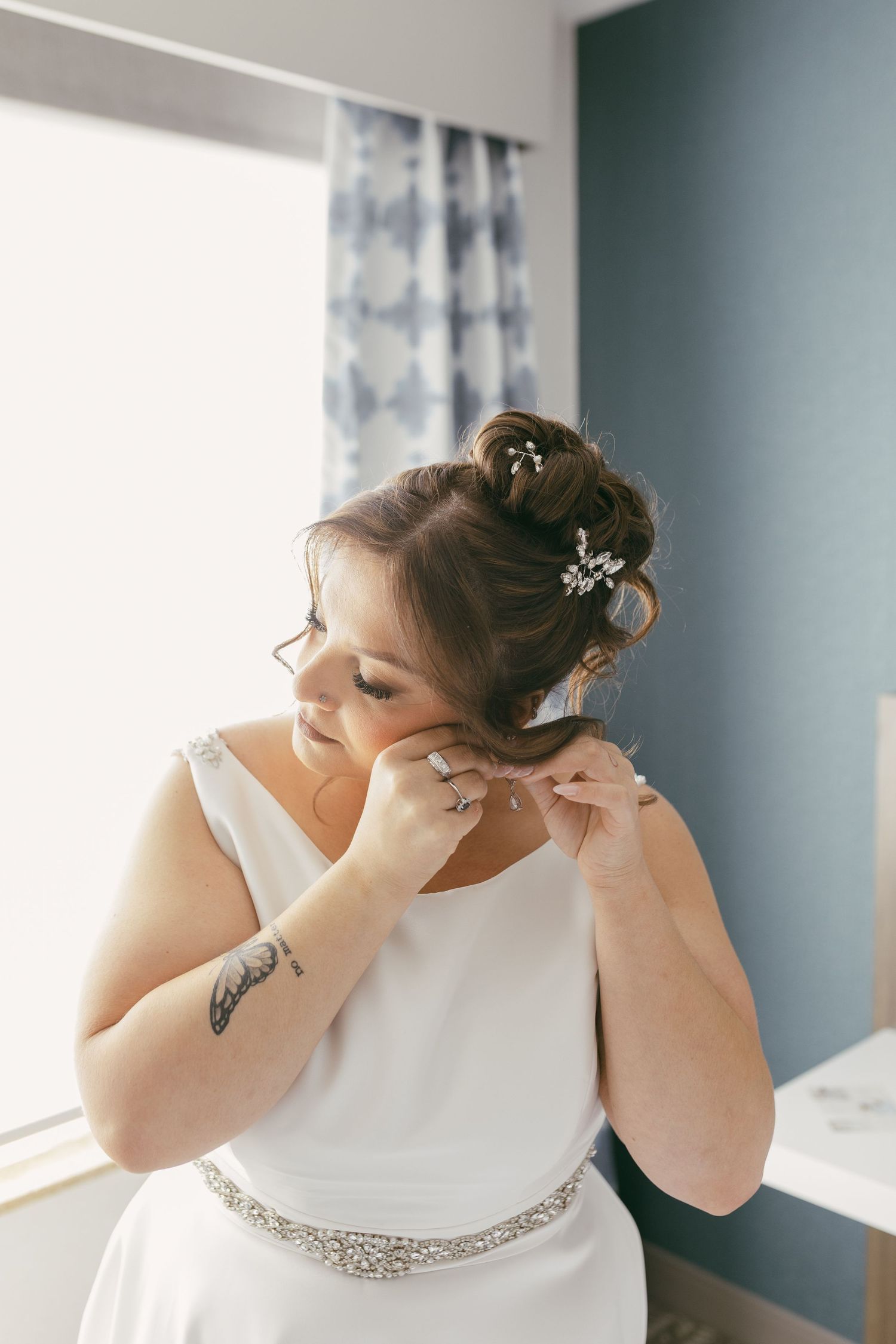 Bride in white gown with crystal belt adjusting earring, hair styled in elegant updo with floral pins.