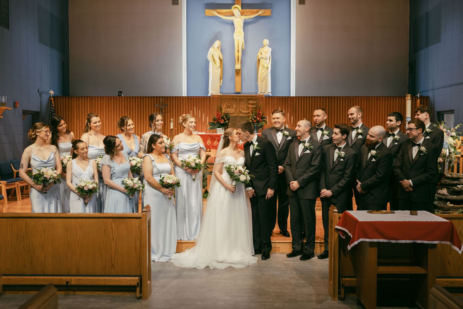 Wedding party poses for a formal photo in a church sanctuary with blue walls and religious artwork.