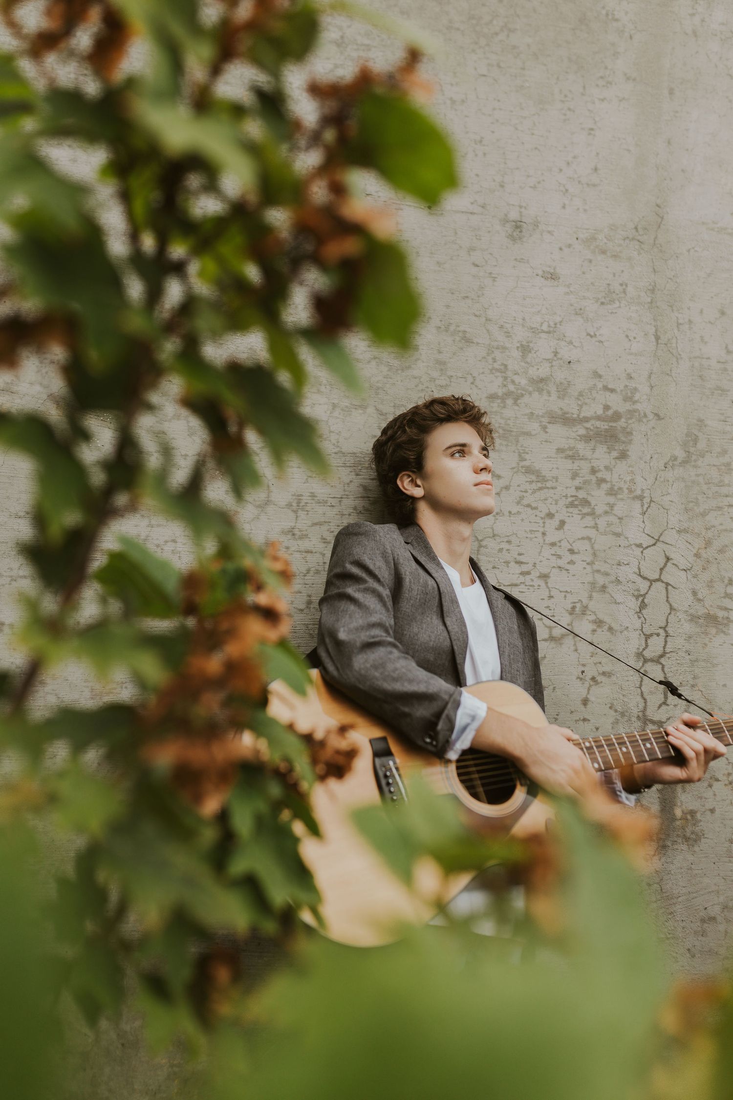 Young man with curly hair plays acoustic guitar outdoors, leaning against a weathered wall surrounded by green foliage.