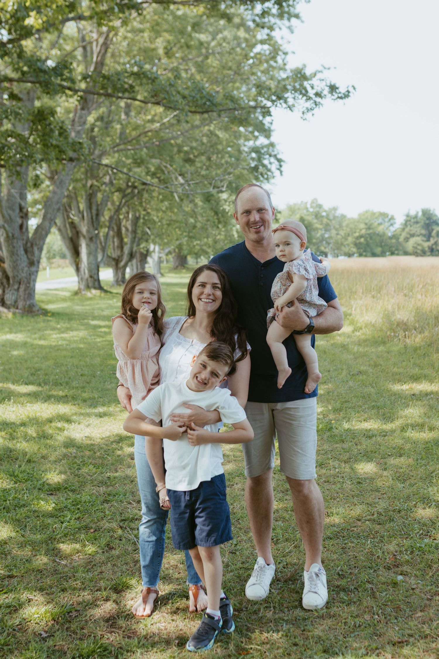 Happy family of five posing outdoors in a lush green park, parents with three young children smiling together.