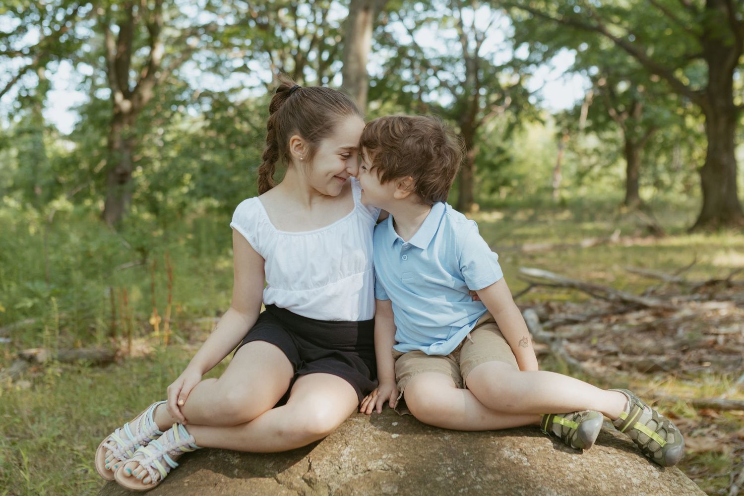 Two children sitting on a rock in a forest, sharing a sweet moment together outdoors.