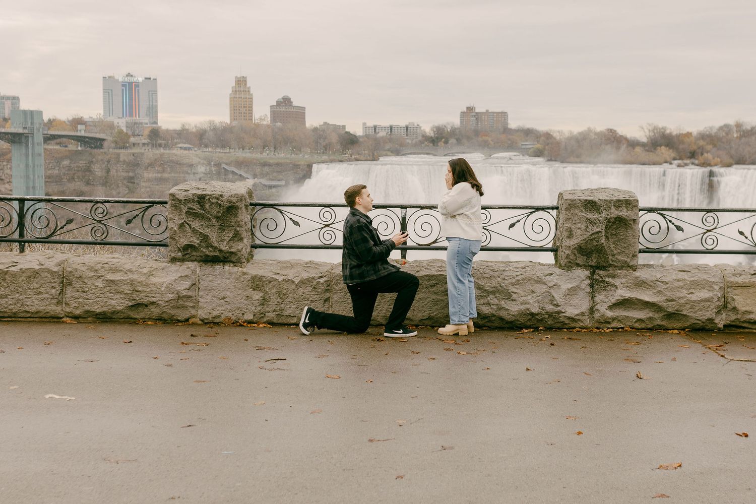 Man proposing on one knee to woman at Niagara Falls overlook with city skyline in background.