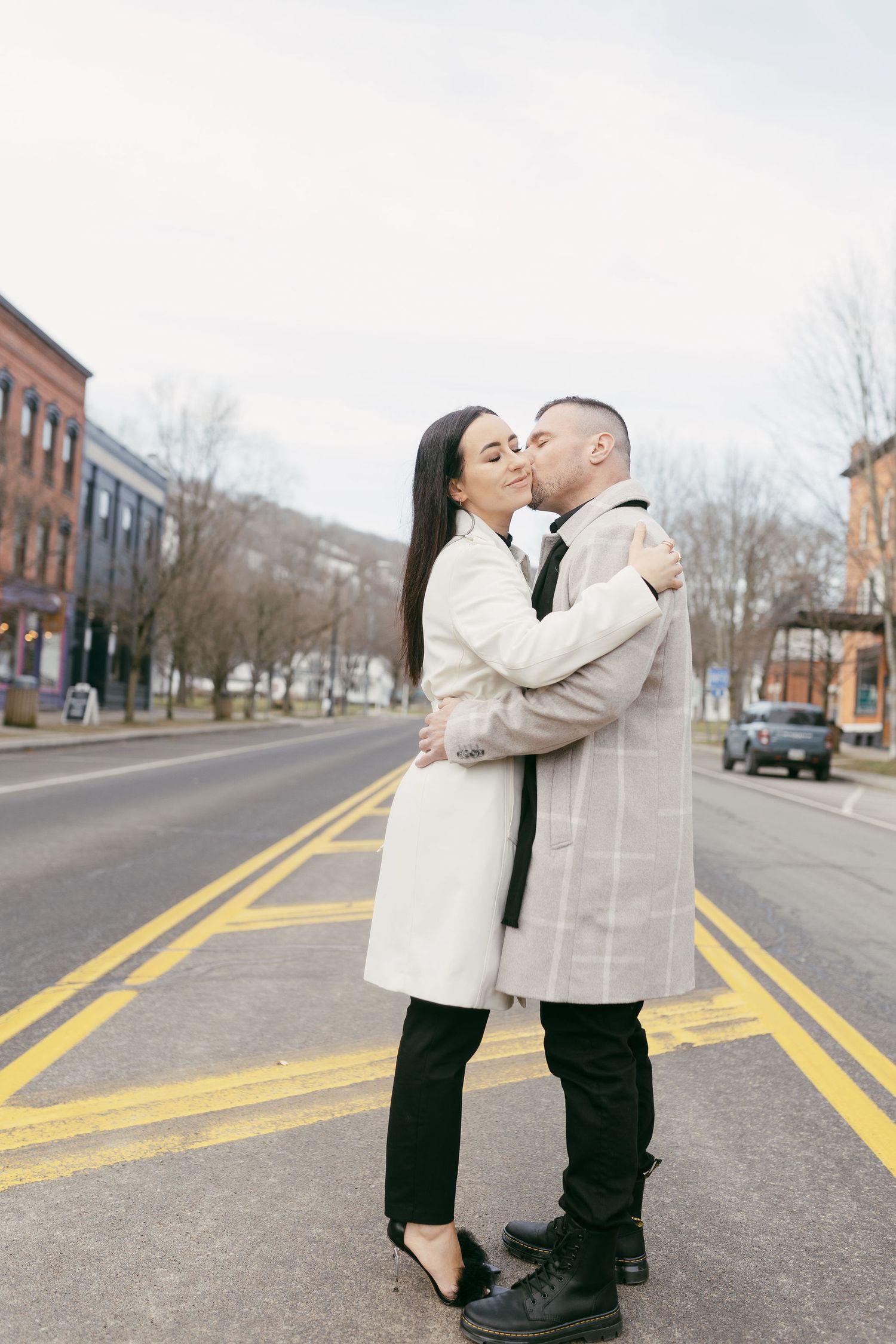 A couple embracing and kissing on a city street, both wearing light-colored coats on a cloudy day.