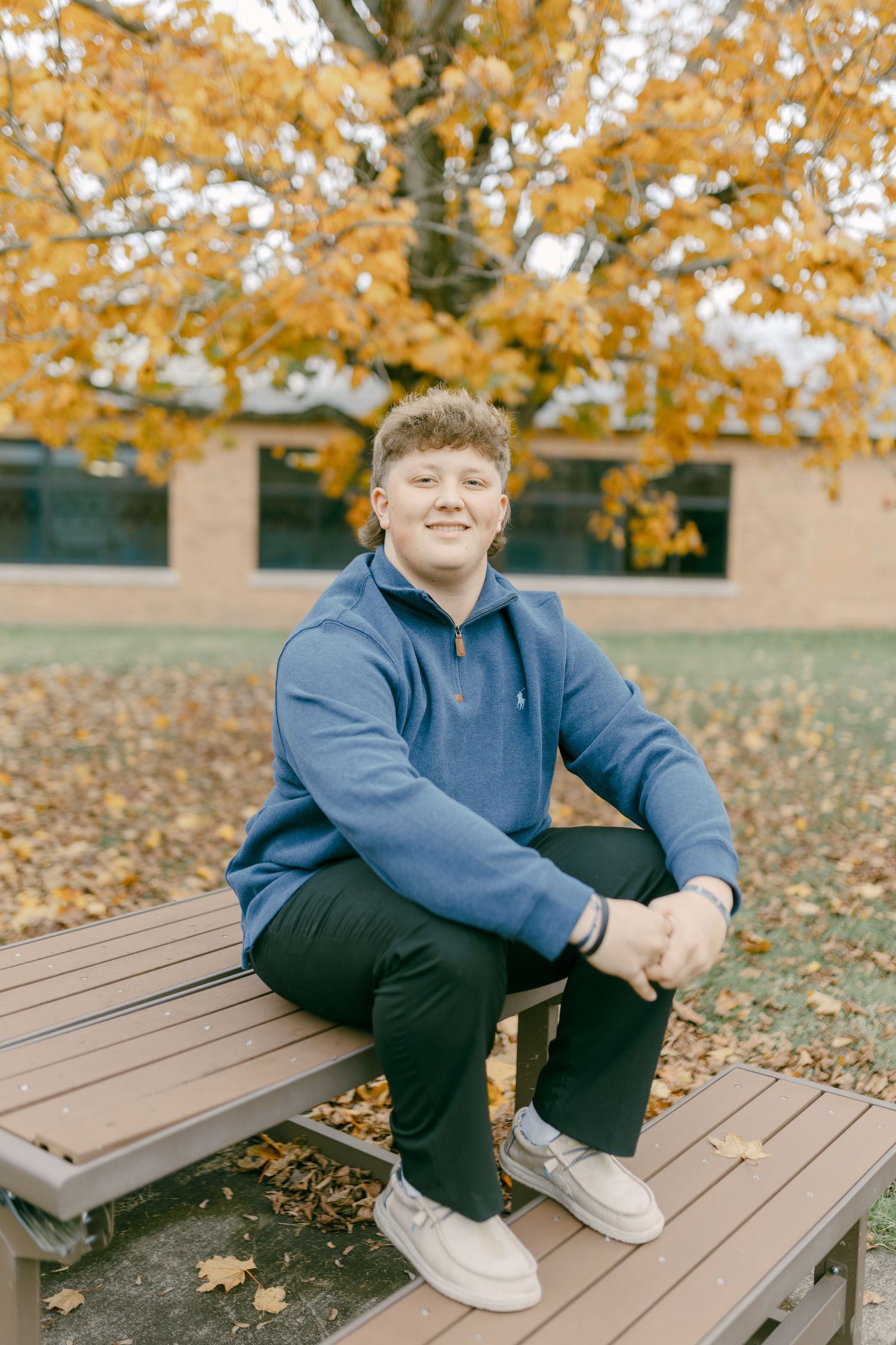 Smiling teen in blue hoodie sits on bench surrounded by golden autumn leaves near brick building.