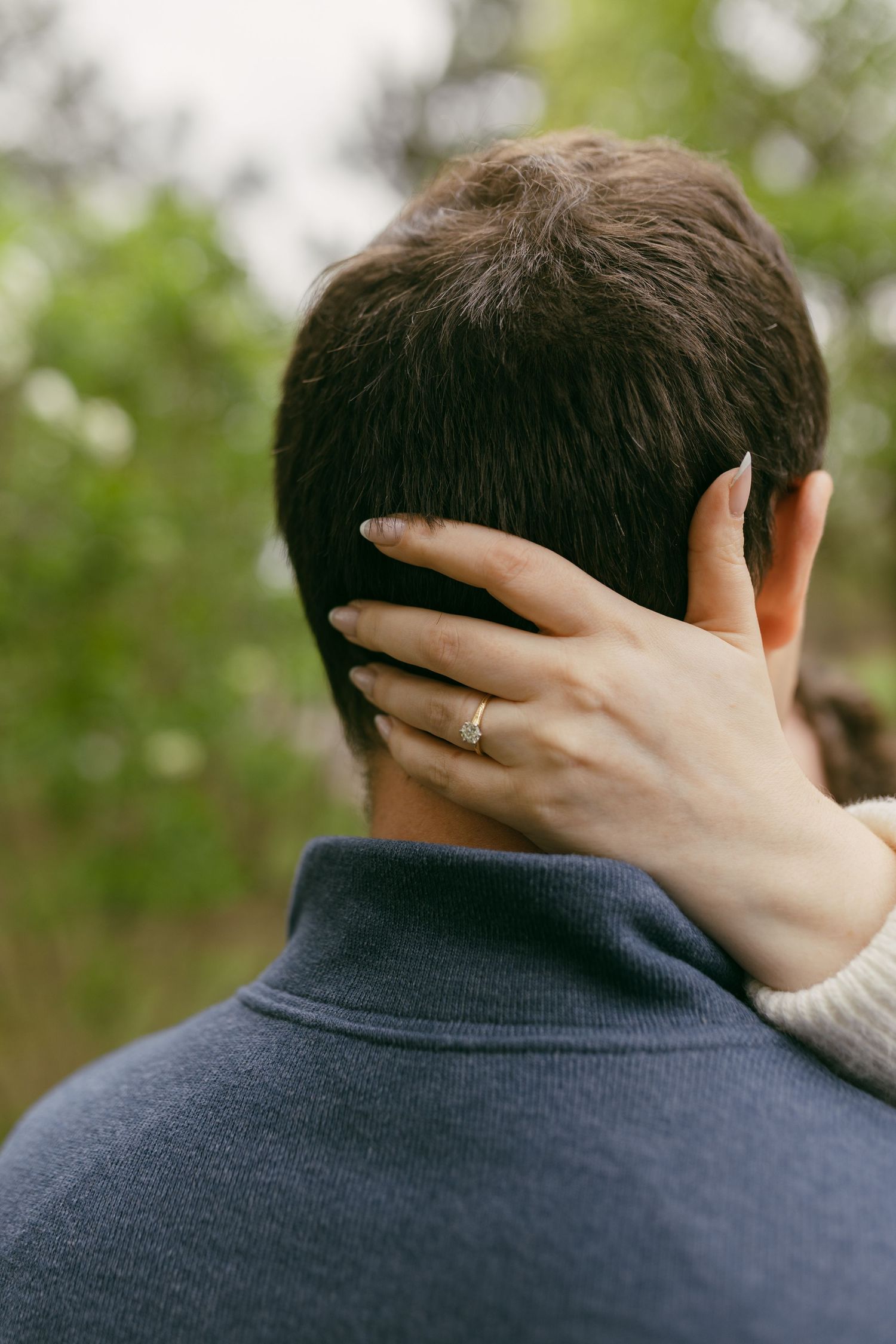 A woman's hand with an engagement ring rests tenderly on the back of a man's neck outdoors.
