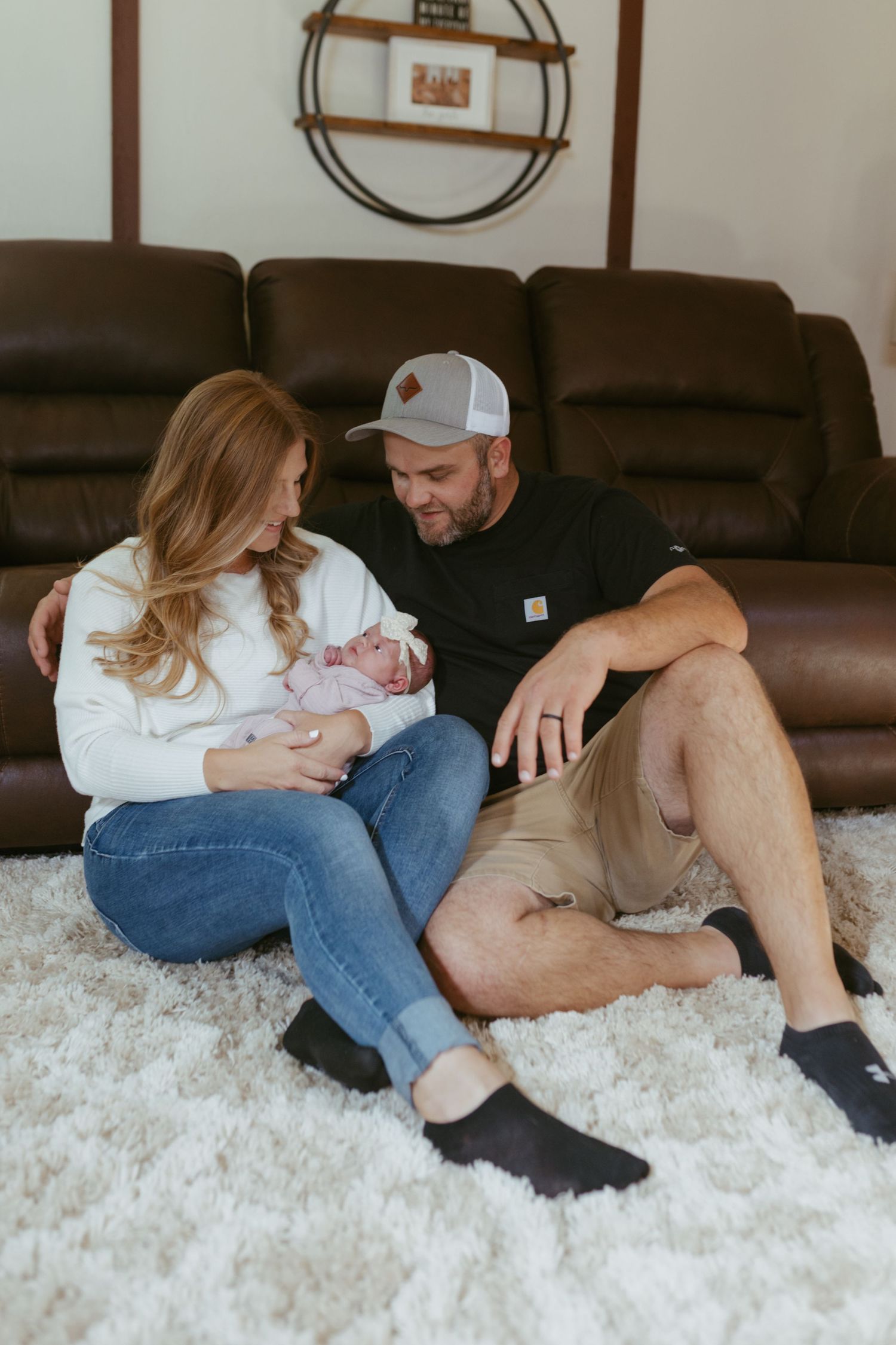 New parents sitting on a white rug holding their newborn baby in front of a brown leather sofa.