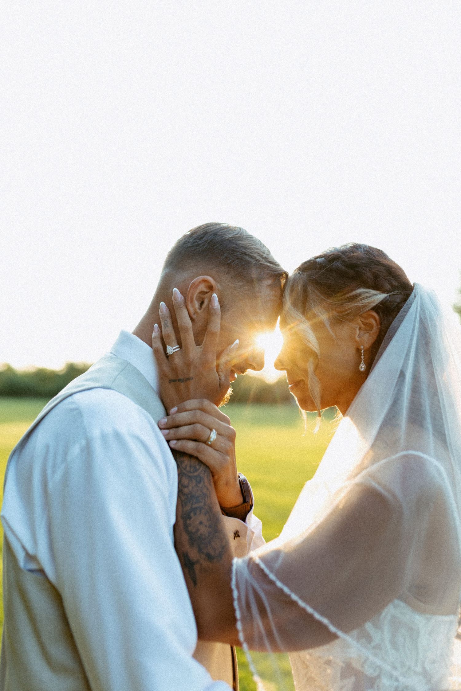 Bride and groom touching foreheads at golden hour sunset, wedding veil and tattoos visible in romantic outdoor portrait.