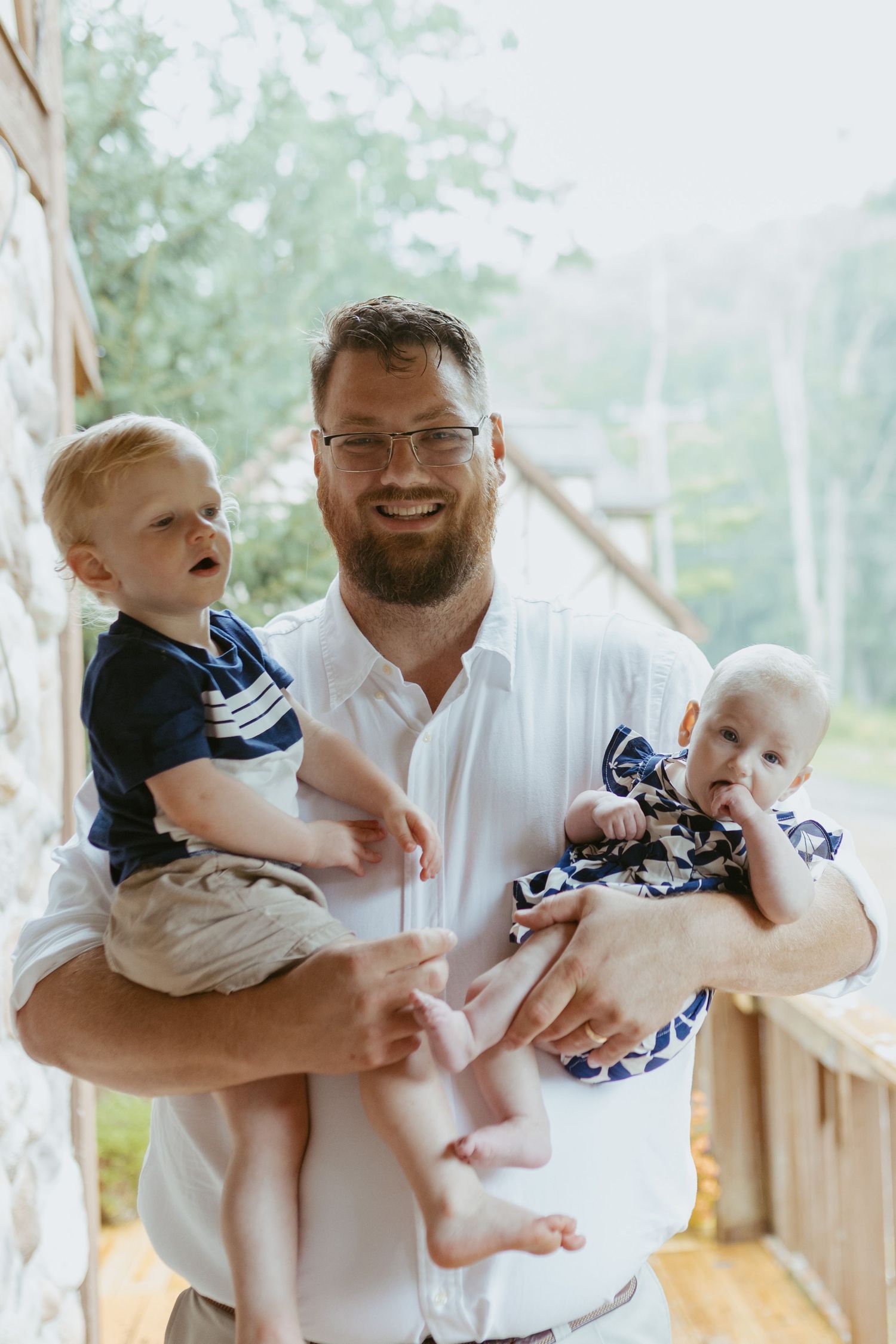 Smiling bearded man with glasses holding two young babies on a wooden porch surrounded by greenery.