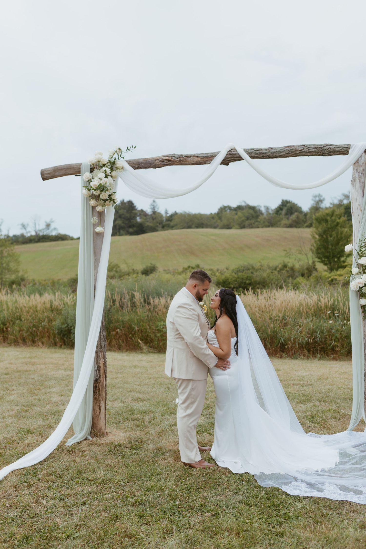 Bride and groom share a romantic moment under a rustic wooden arch draped with white fabric in a scenic outdoor setting.