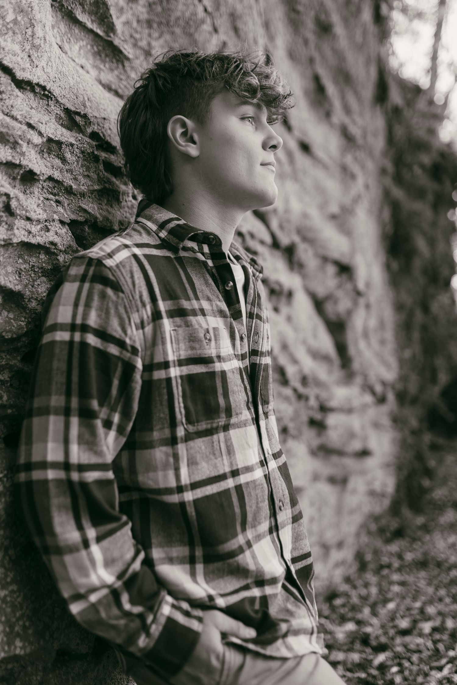 Black and white portrait of a teenage boy in plaid shirt leaning against a stone wall, looking upward thoughtfully.
