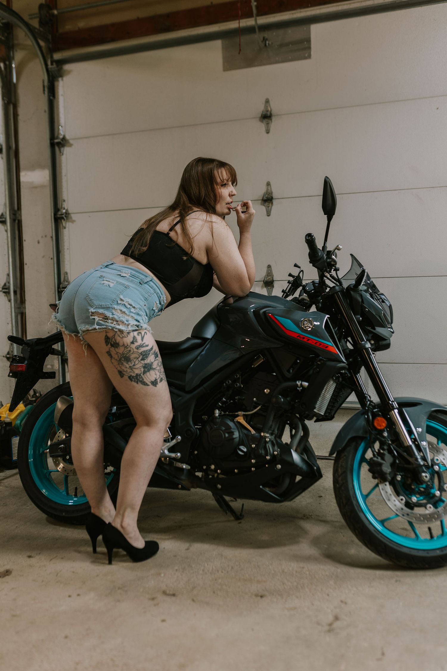 Person in casual attire posing with dark motorcycle in garage setting.